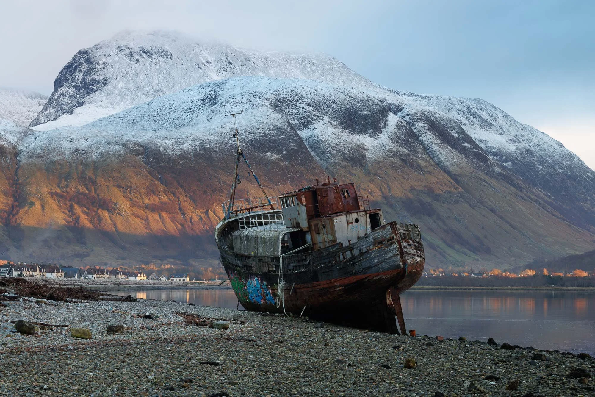 An abandoned, rusted shipwreck resting on a rocky shoreline with a Ben Nevis Scotland in the background.