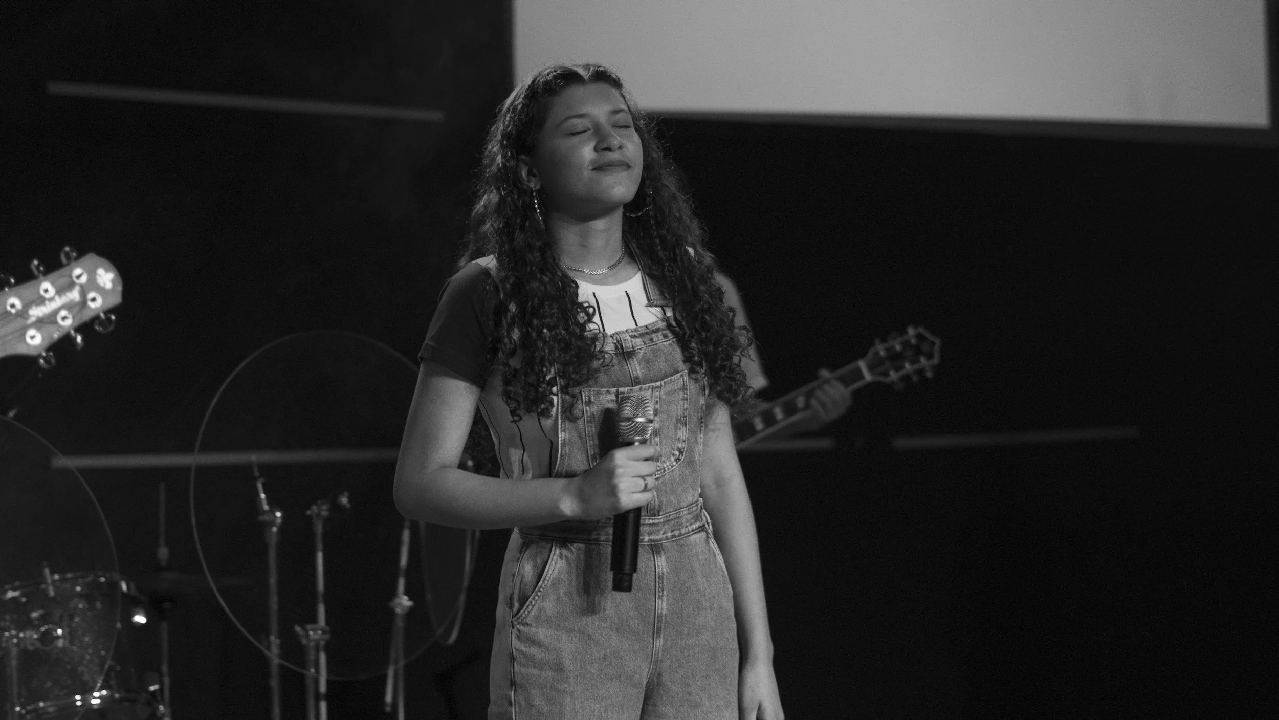 A young woman with curly hair holding a microphone with her eyes closed, standing on stage with musical instruments in the background.