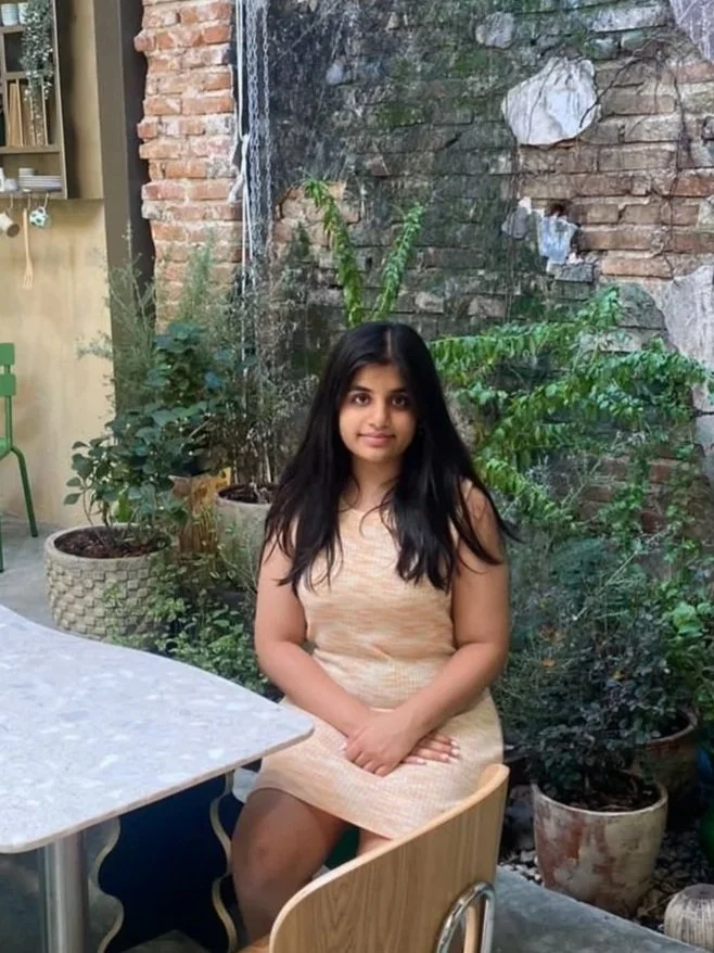 Young girl with long dark hair sitting at a table in a garden setting with green plants and a brick wall background.