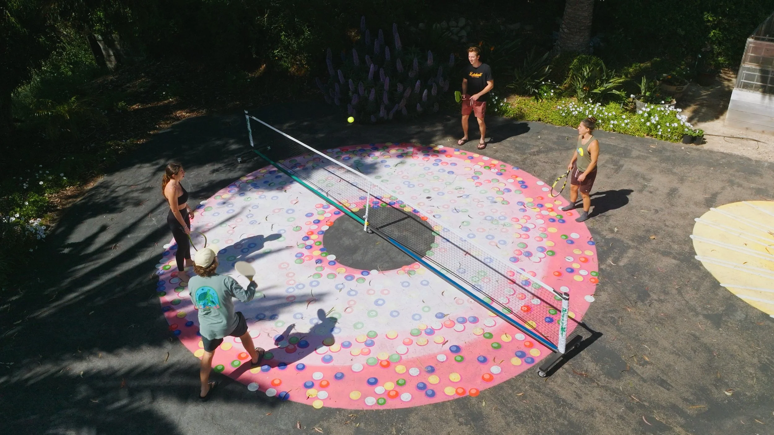 People playing a game of tennis with a ball and paddles on a circular court decorated with colorful dots, surrounded by trees and plants.