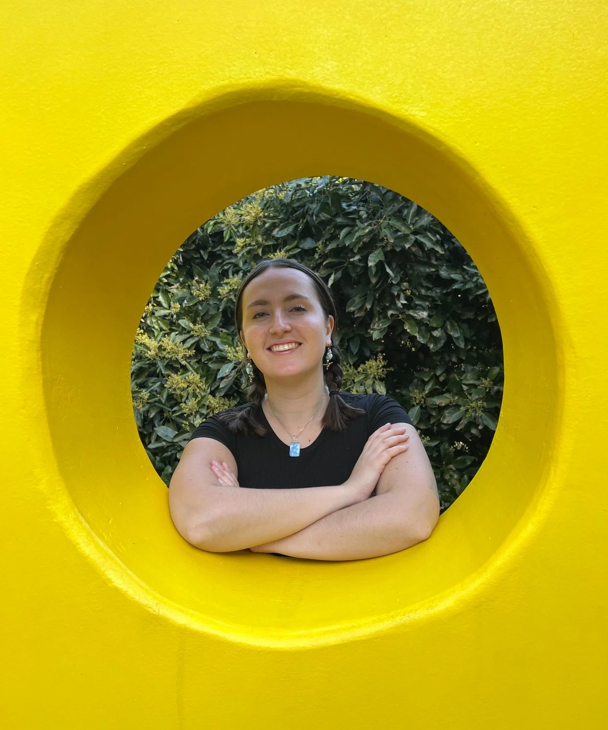 A woman with dark hair and fair skin smiling, wearing a black top with jewelry, crossing her arms, standing behind a yellow circular frame with a background of green foliage.