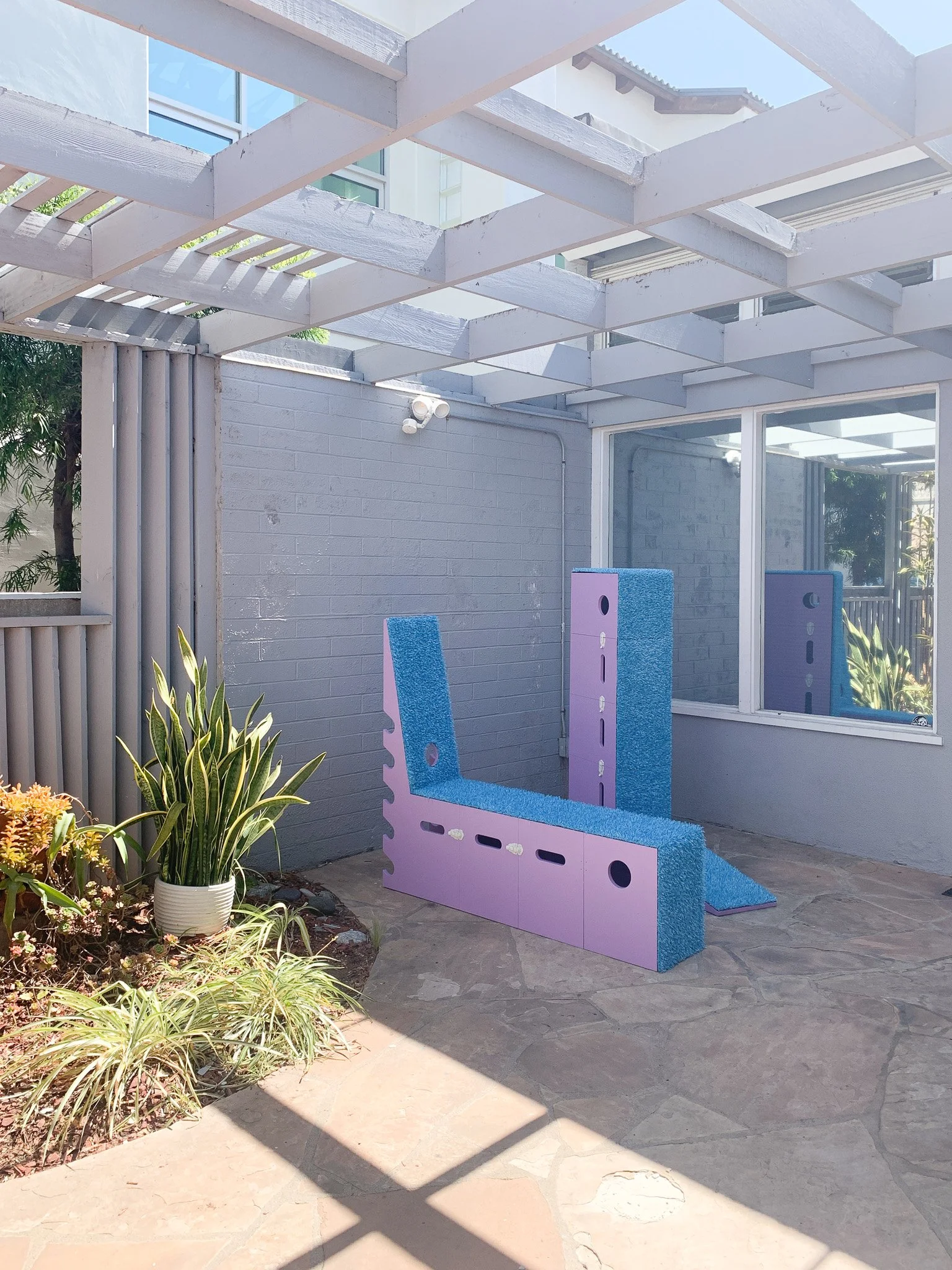 Outdoor patio with a gray brick wall, potted plants, and a purple and blue climbing structure for children under a pergola.
