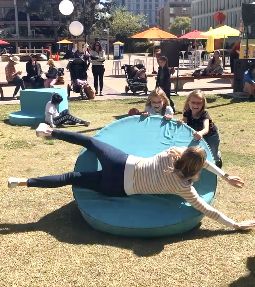 Two women sitting on large colorful outdoor chairs, surrounded by children playing on a grassy area with trees and buildings in the background on a sunny day.
