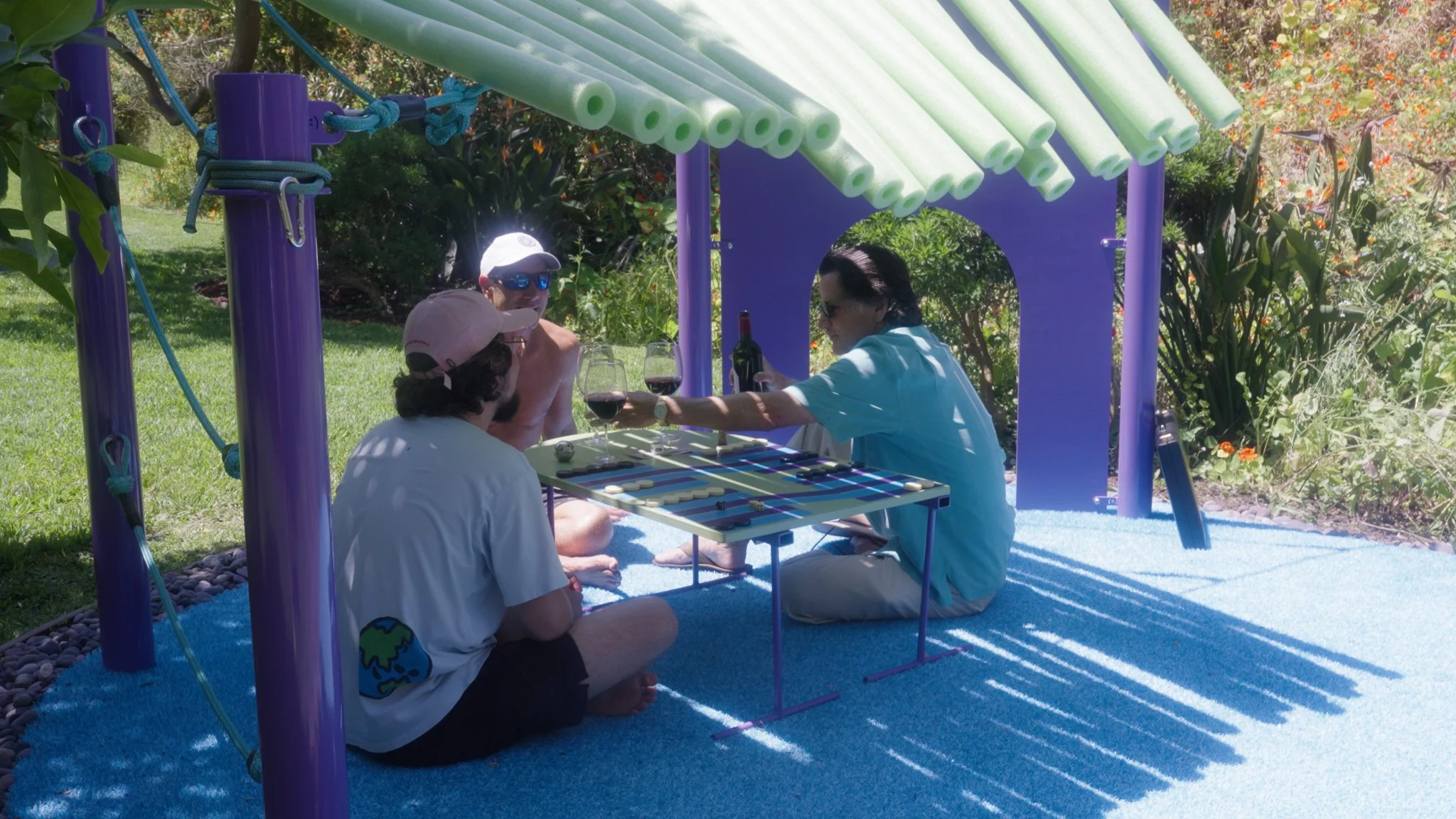 Three men playing a game of backgammon outdoors under a small purple and green playhouse structure, with two holding wine glasses and one pouring wine.