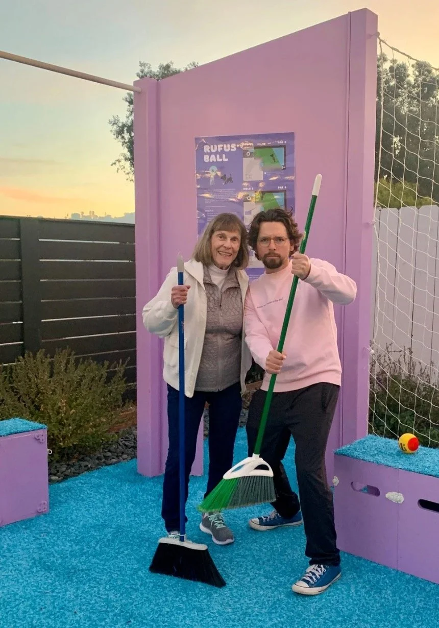 Two people holding broomsticks, standing on a blue textured surface with a purple backdrop, possibly outdoors at sunset.