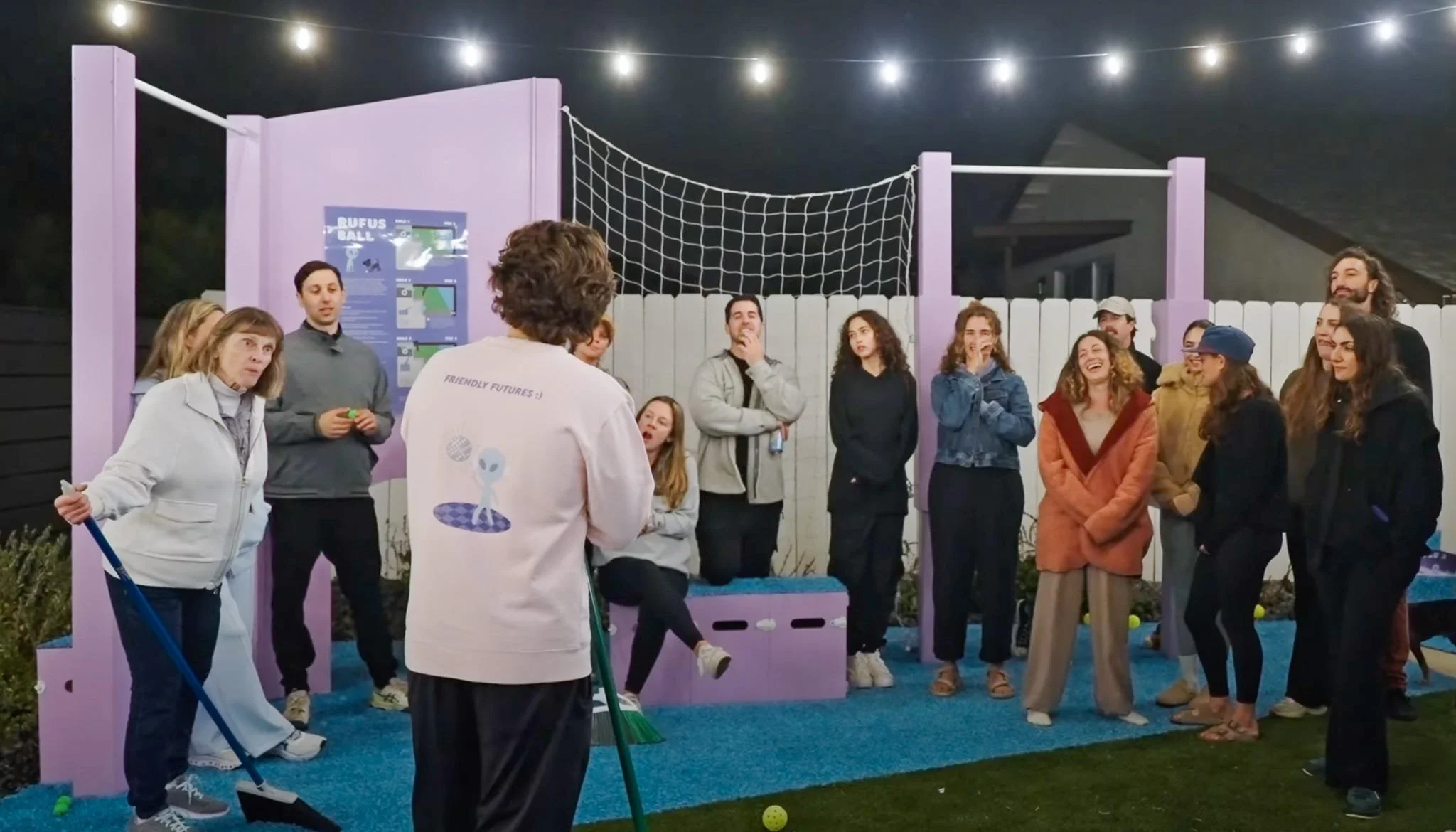 A group of people gathered outdoors in front of a purple wall with a volleyball net. They are listening to a speaker, with some smiling and laughing. There are string lights overhead, a few yellow tennis balls on the ground, and a poster on the wall.