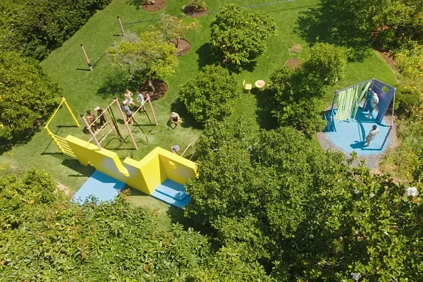 A playground with swings, a yellow slide, a blue climbing structure, and children playing surrounded by green trees and grass.