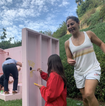 People painting a pink wooden structure outdoors, with a woman in a white tank top and shorts smiling, and a girl in a red jacket painting near the top.