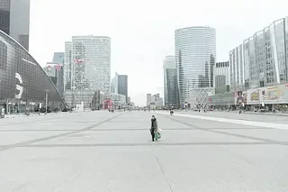 A lone person standing in an empty urban plaza surrounded by modern high-rise buildings.