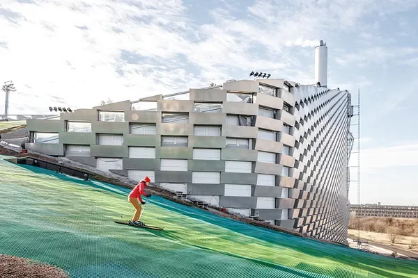 A person snowboarding on a green artificial slope in front of a modern white building with unique architectural design.