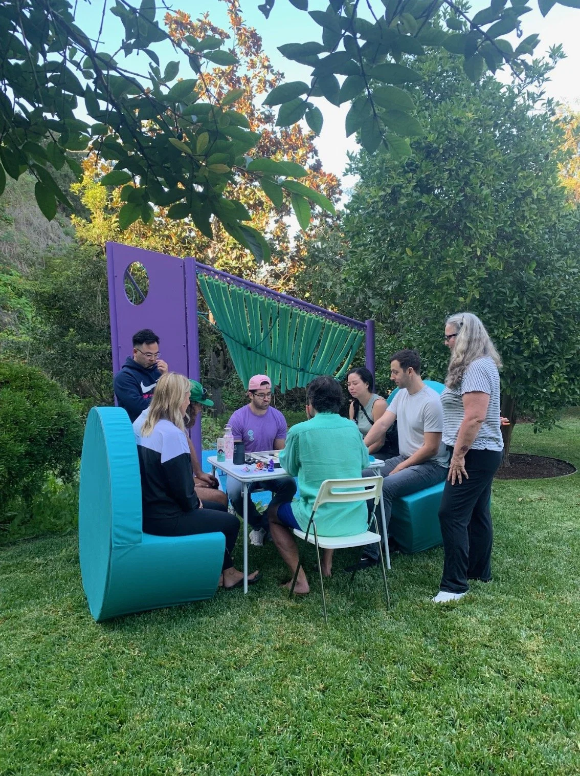 A group of seven people gathered outside around a table in a backyard, with colorful seating and a purple playground structure with green ropes in the background.