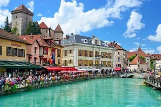 Bright daytime view of a European riverside with colorful buildings, outdoor cafe seating, and a castle with a tower in the background.