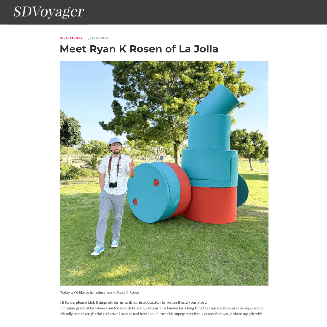 A man with glasses, a beard, wearing a white short-sleeve shirt, a hat, and a camera around his neck, stands outdoors in front of a large art installation consisting of colorful cylindrical shapes. Behind him is a tall green tree and a park with grass and other trees.
