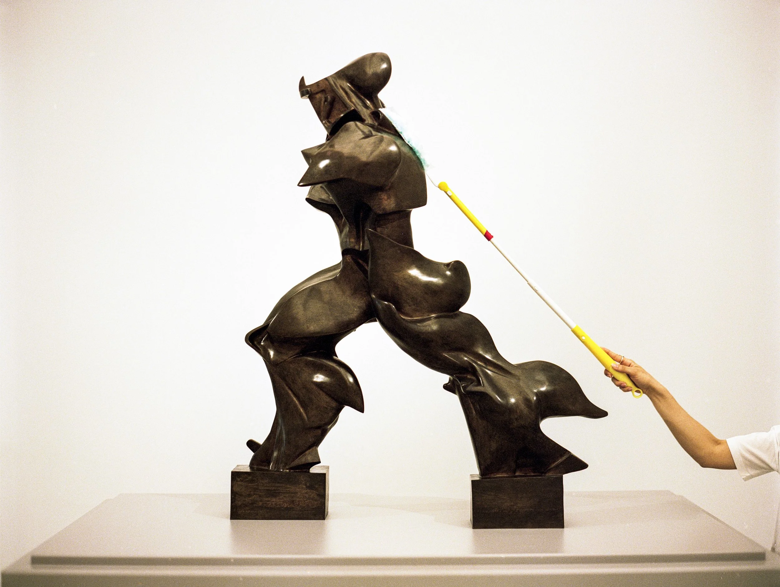 An abstract bronze sculpture of a dog with textured, rounded forms, placed on a pedestal, with a person using a long cleaning tool to wipe it.