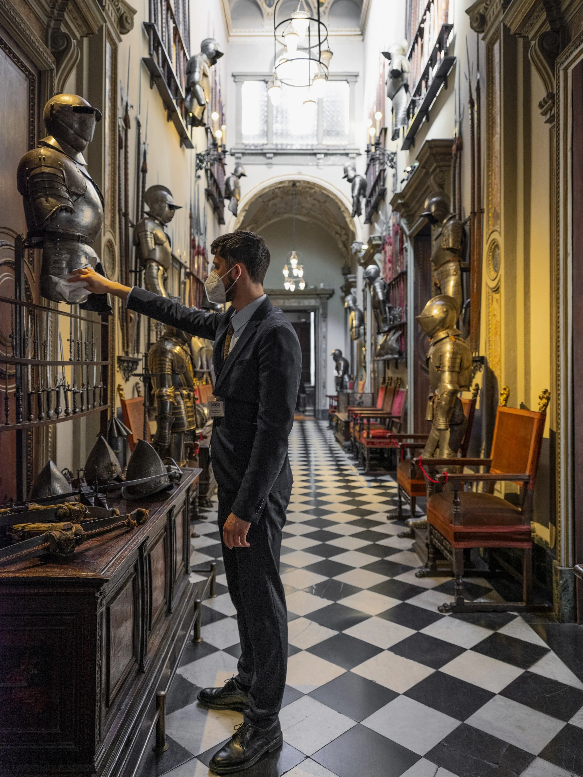 A man wearing a suit and a face mask is polishing a suit of armor in a historic museum hallway filled with medieval armor displays, red velvet chairs, and checkered black and white flooring.