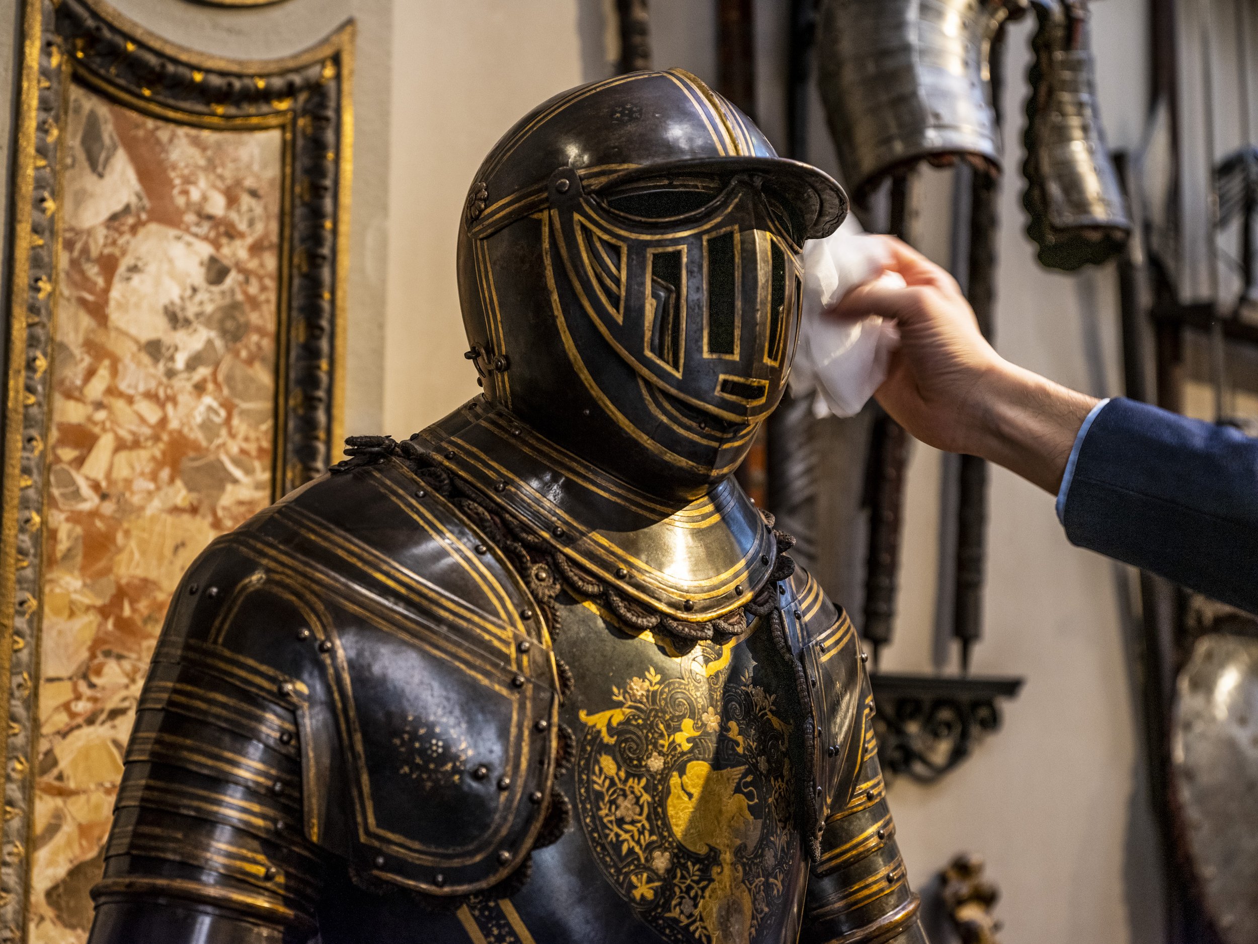 Person cleaning a suit of black and gold medieval knight armor with ornate designs, in a museum setting.