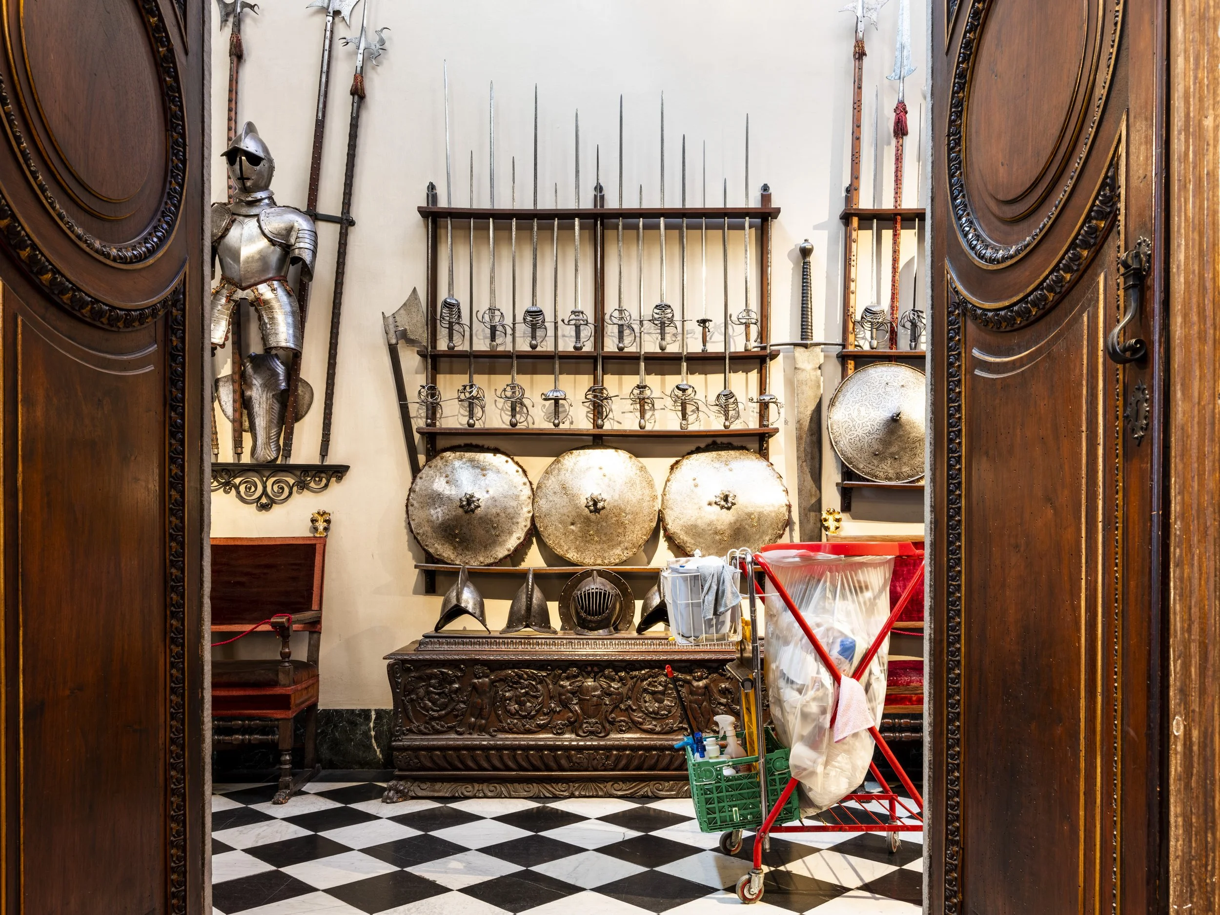 Room with medieval armor, swords, shields, and helmets on display, viewed through wooden doorway with checkered black and white floor.