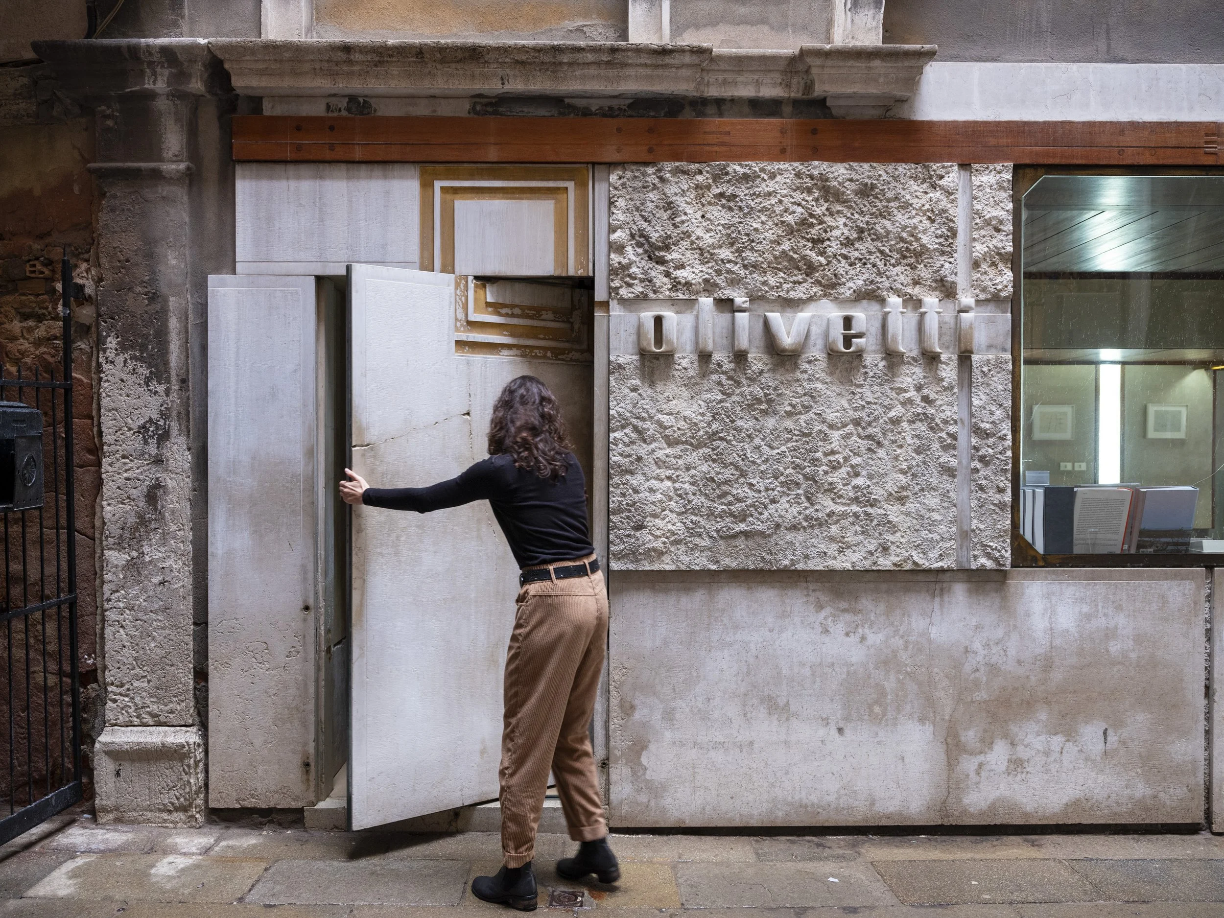 A woman with curly hair, dressed in a black top and light brown pants, opening a door with her left hand in front of a stone wall with the word 'Olive' spelled out in large letters.
