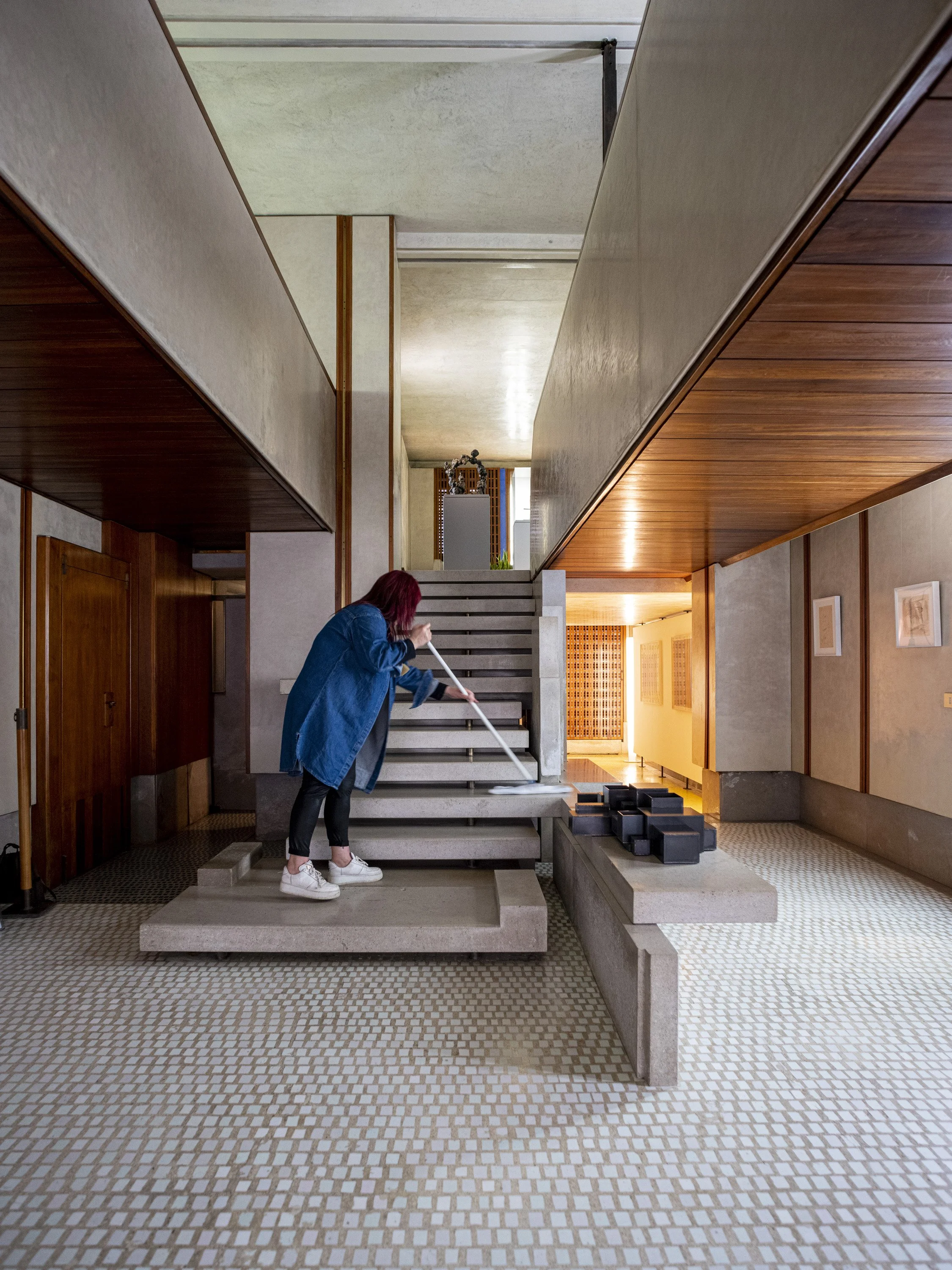 A woman with red hair cleaning a staircase with a mop in a modern, minimalistic interior space with wood-paneled ceiling and framed pictures on the wall.