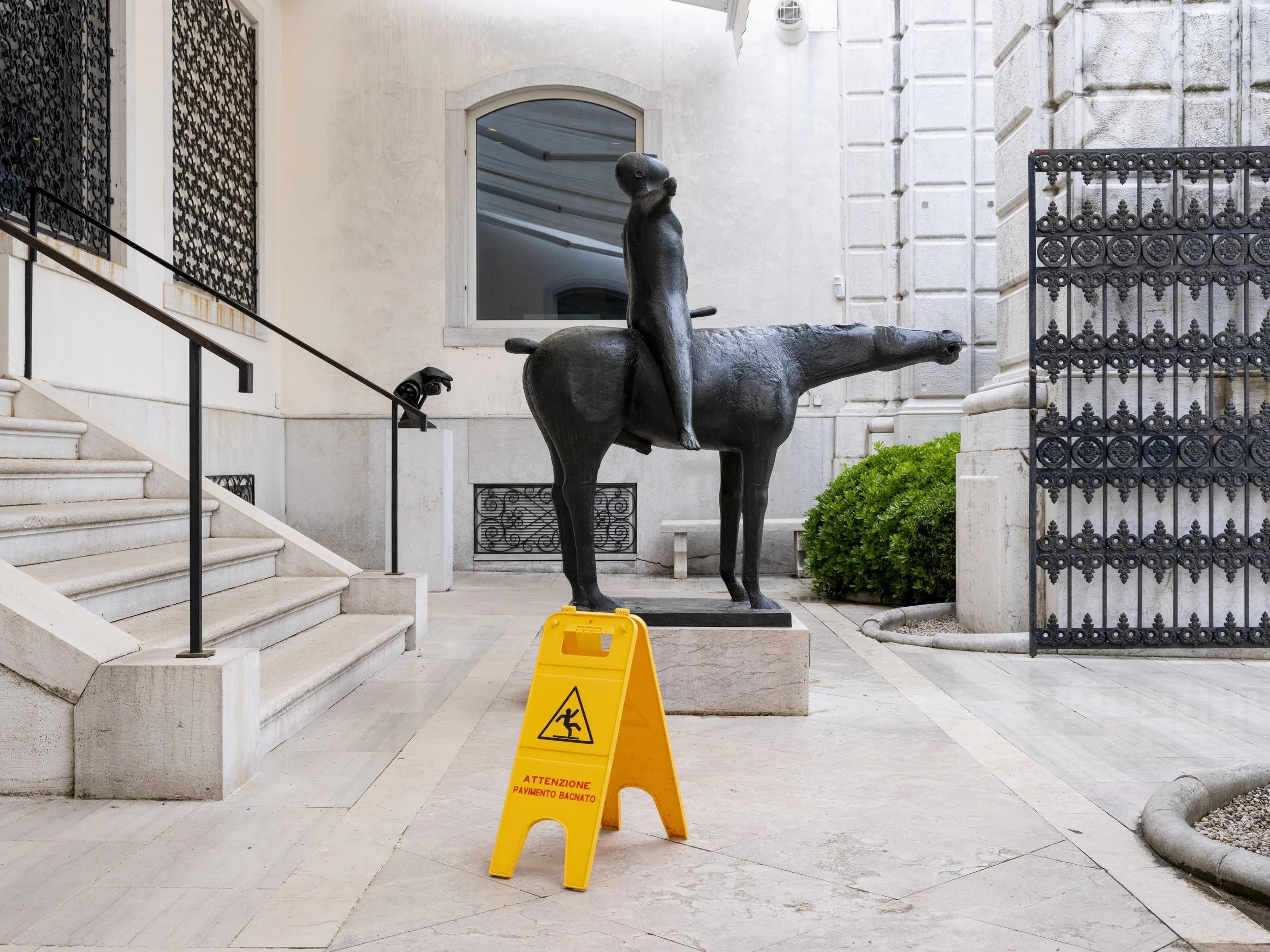 A black sculpture of a creature with the body of a horse and the head and torso of a humanoid figure, situated outdoors near white stone stairs and a building wall. A yellow caution sign warning of slippery pavement is in the foreground.