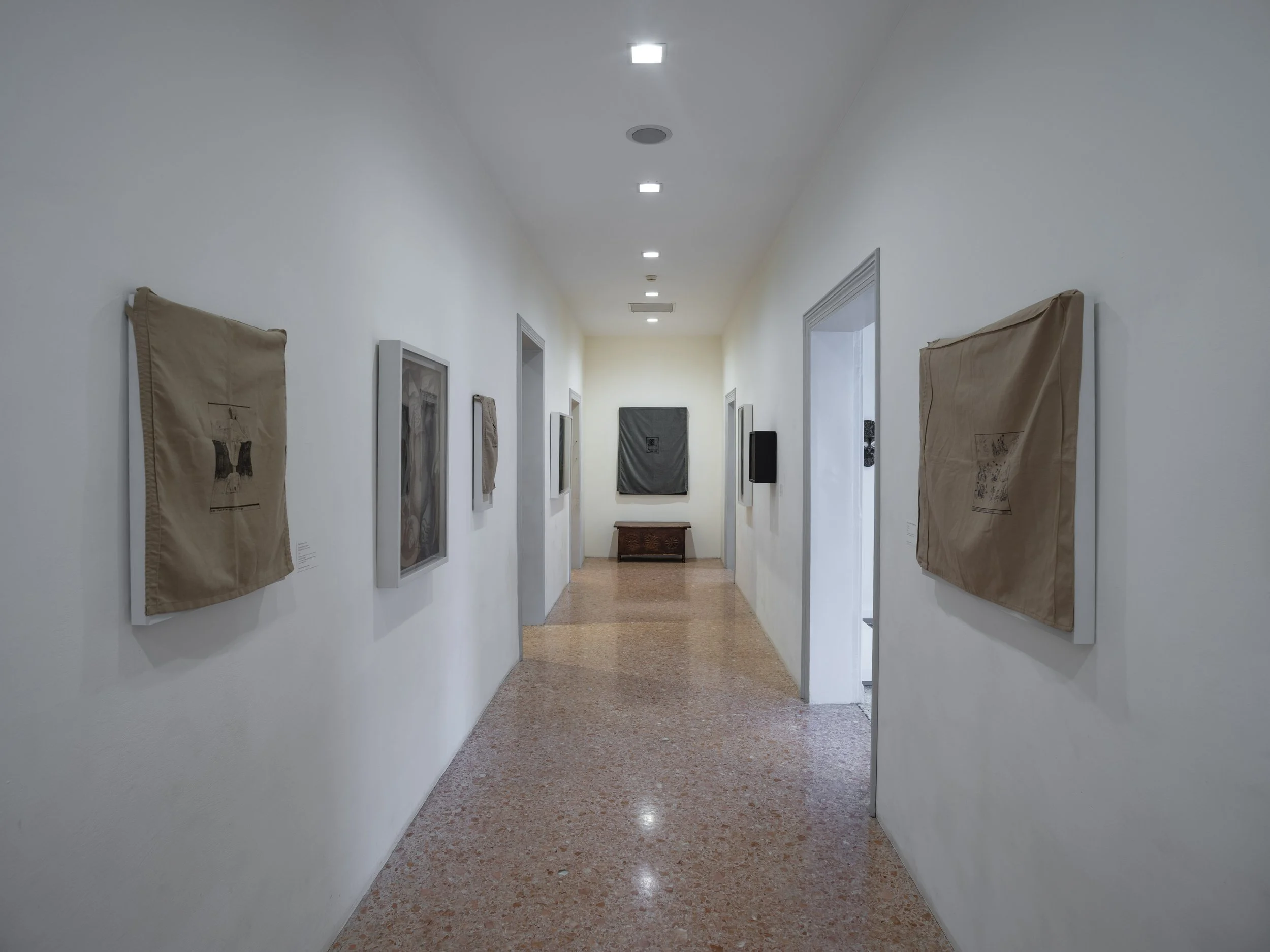 An art gallery hallway with framed artworks on white walls and a small bench at the end.