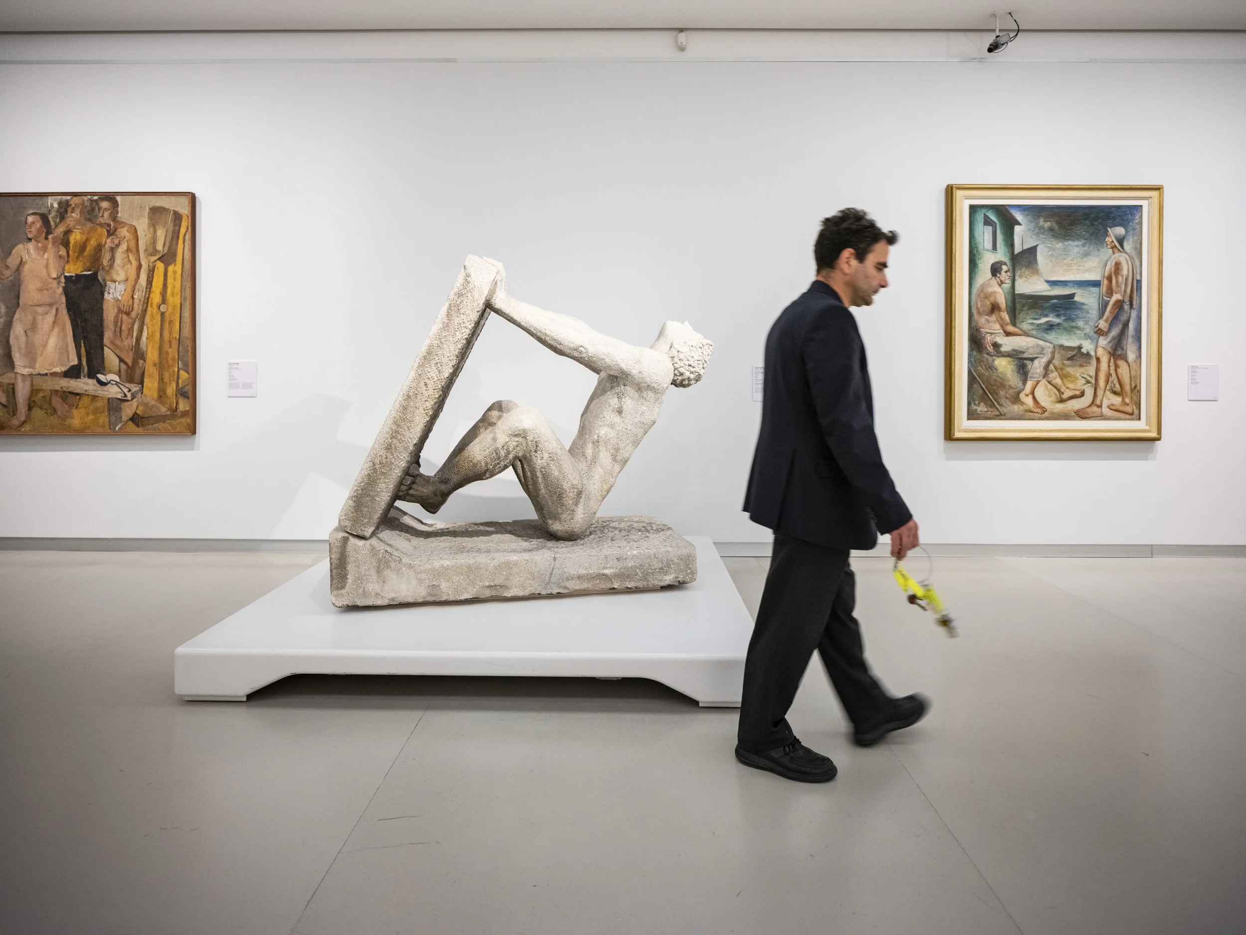 Man walking past modern sculpture of a reclining figure holding a large rectangular object in an art gallery with framed paintings on white walls.