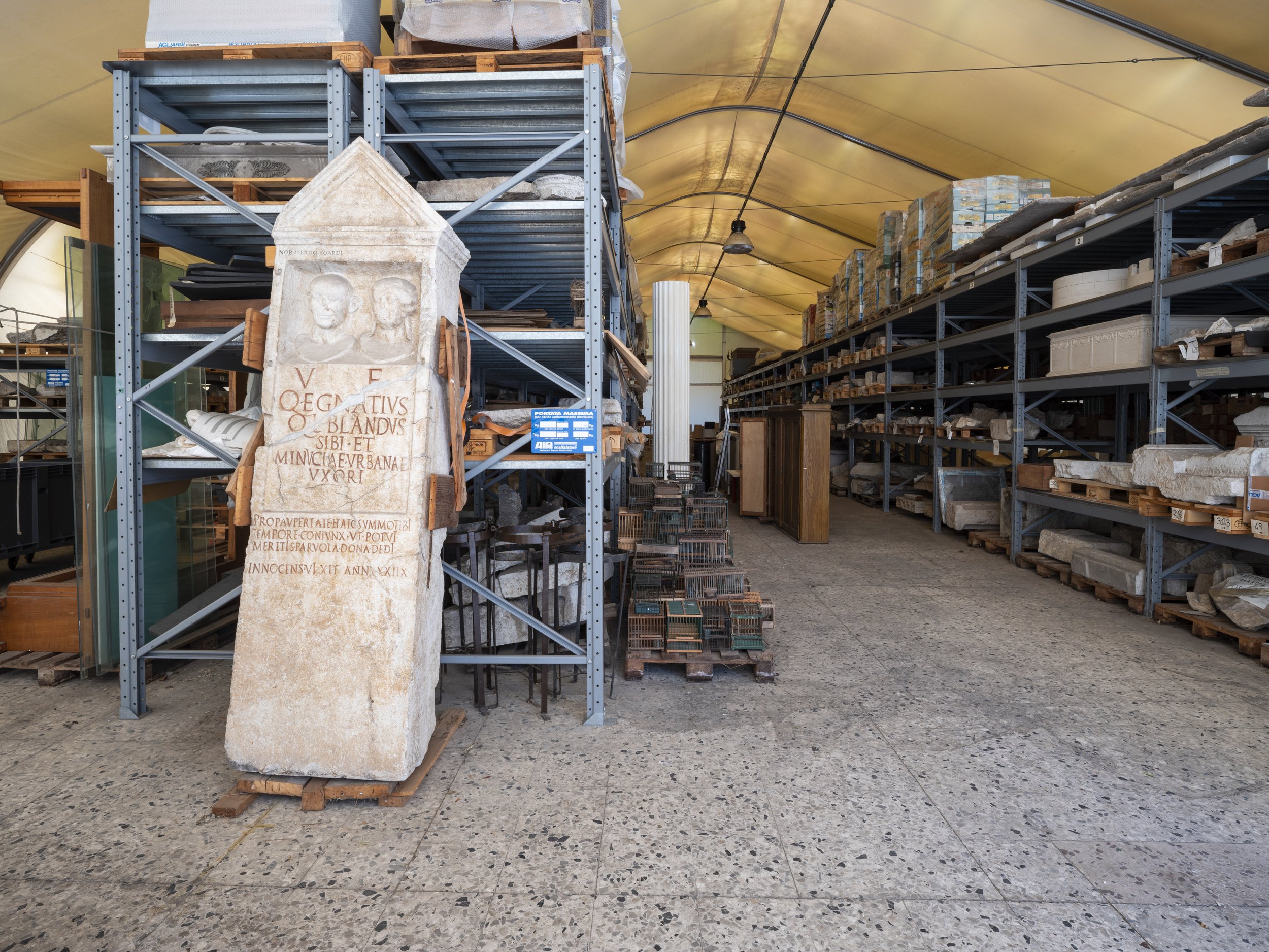 A storage warehouse with shelves filled with stone artifacts, statues, and architectural fragments. In the foreground, there is a tall stone slab with Latin inscriptions and a carved relief at the top. The warehouse has a yellow arched ceiling and a 