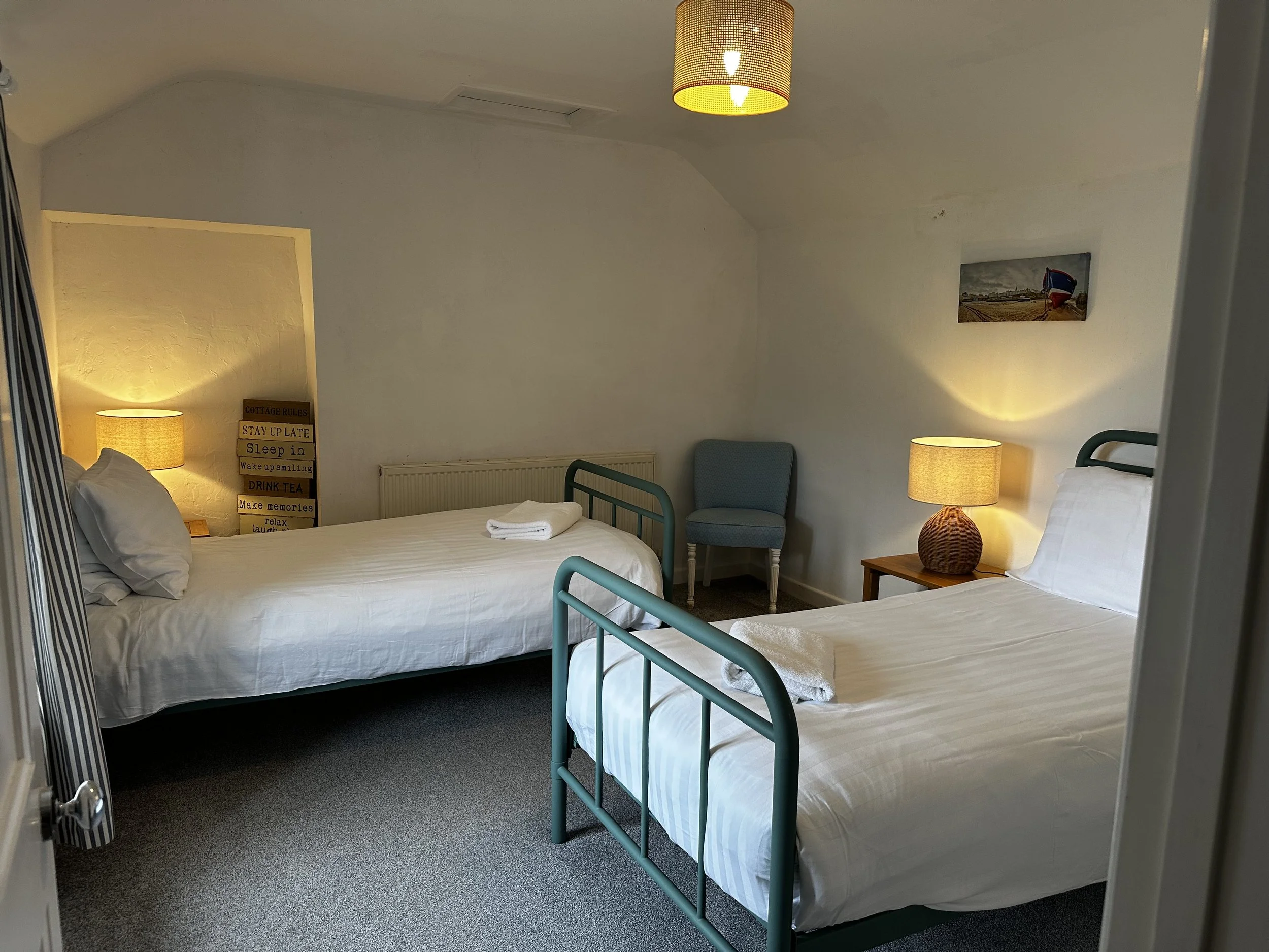 Bedroom with two single beds, white linens, a wooden nightstand with a lamp, a blue armchair, and a decorative sign on the wall with stay-up-late rules.