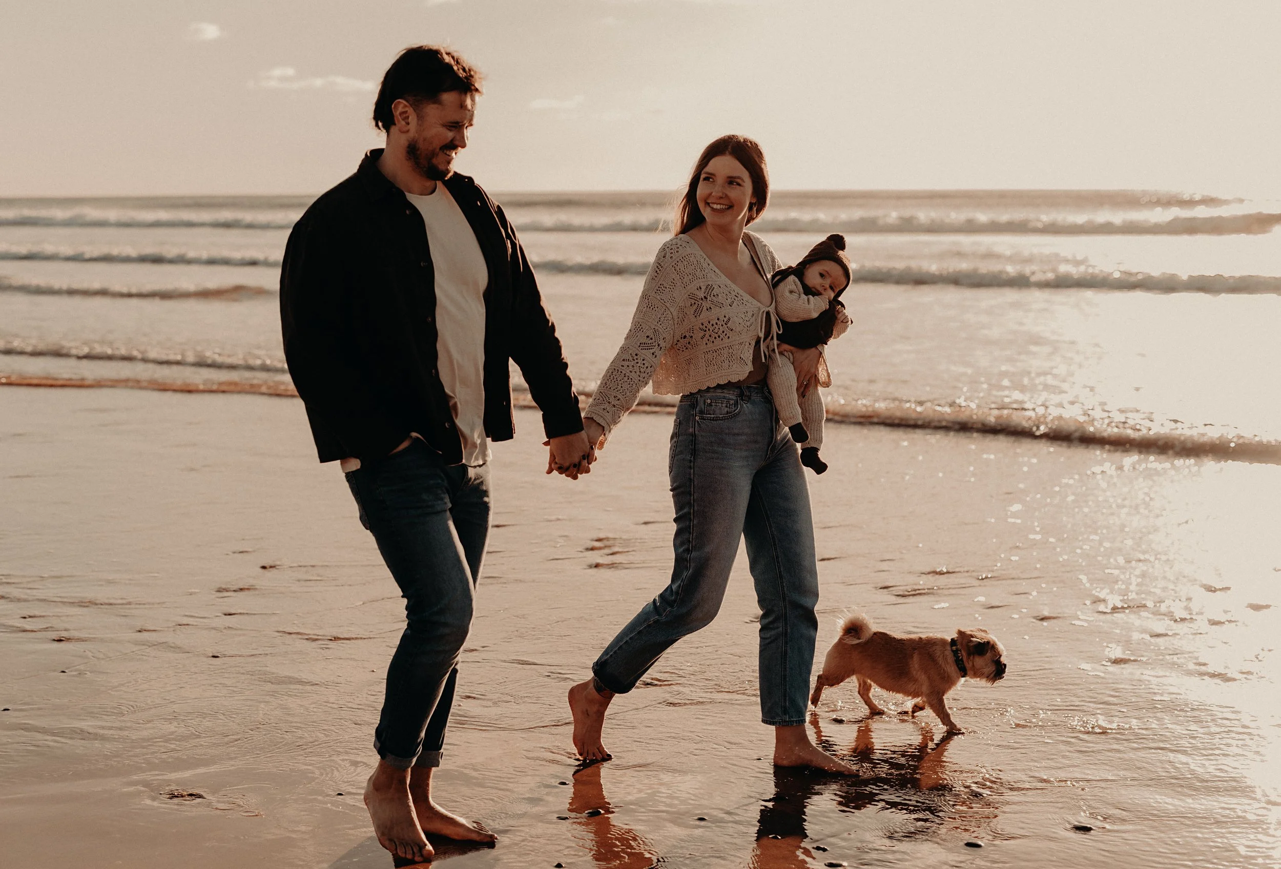 A family of three walking barefoot on the beach at sunset, holding hands with their dog, with the ocean in the background.