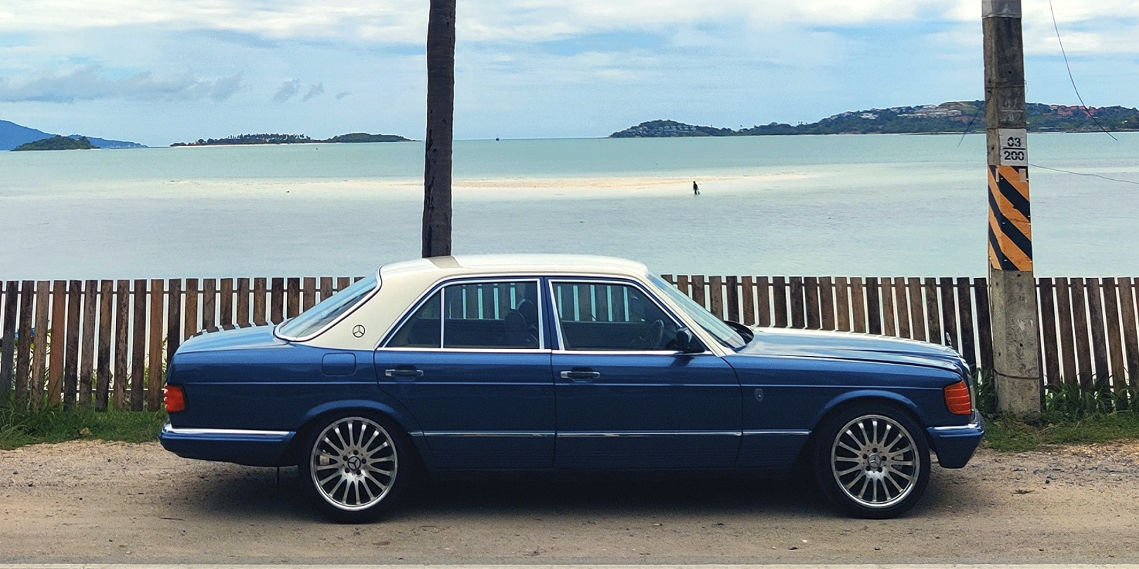 Vintage blue Mercedes-Benz parked beside a wooden fence near a beach with calm water, small islands, and a cloudy sky in the background.