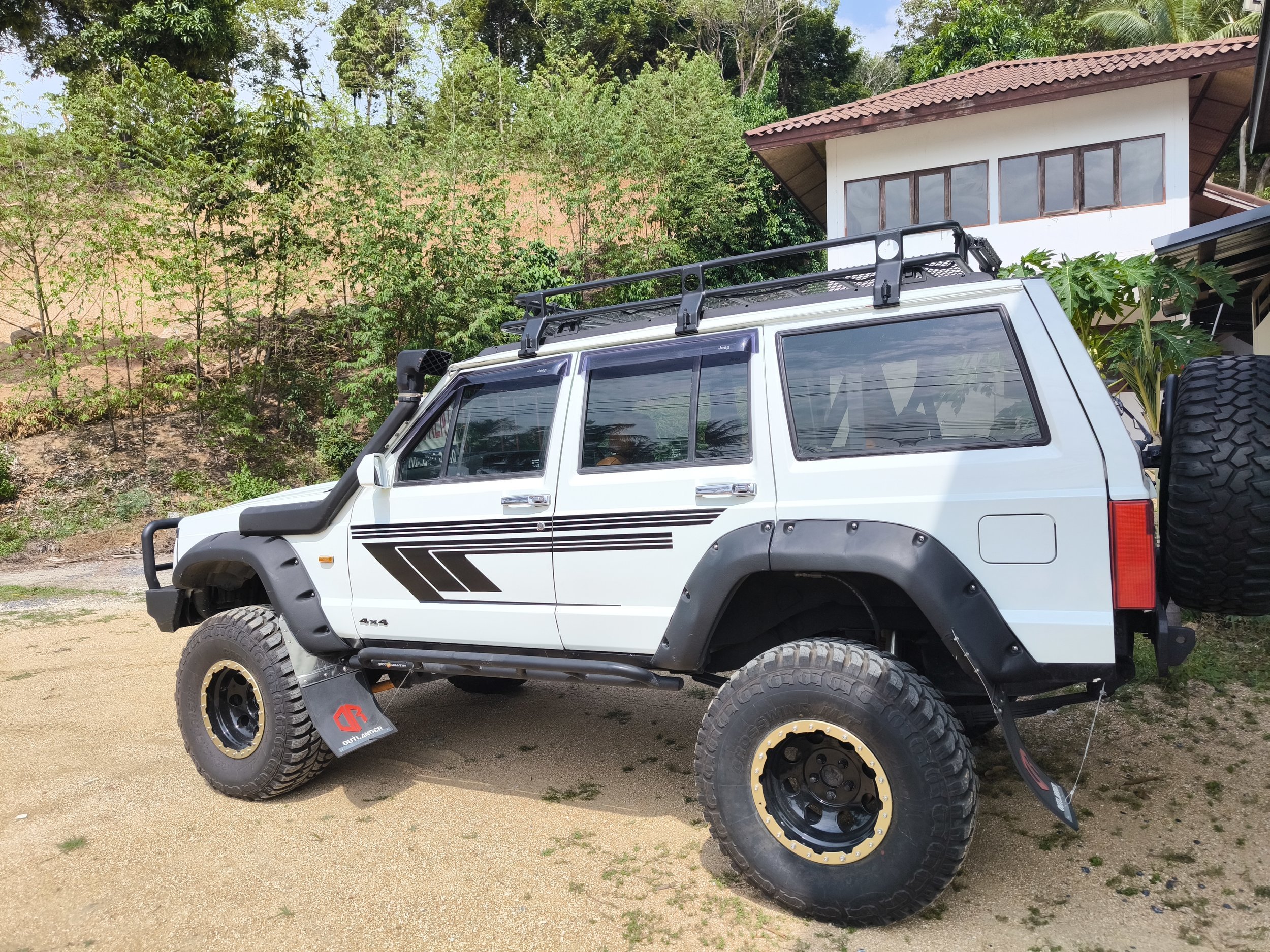 A white off-road Jeep vehicle parked on a dirt surface in front of a house with a red-tiled roof, surrounded by lush green trees and plants.