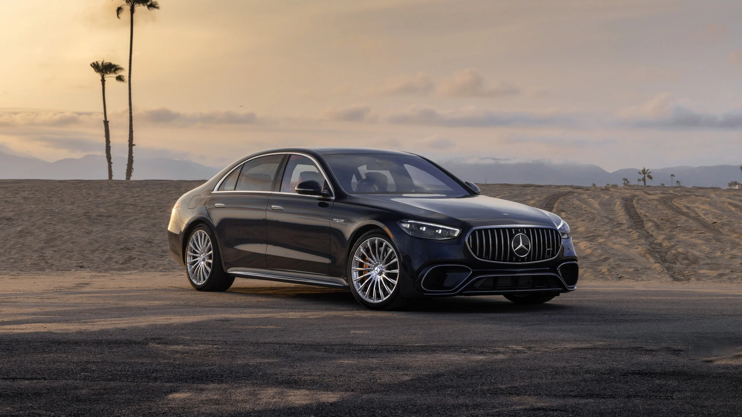 A black Mercedes-Benz luxury sedan parked on a sandy road with palm trees and desert mountains in the background during sunset.