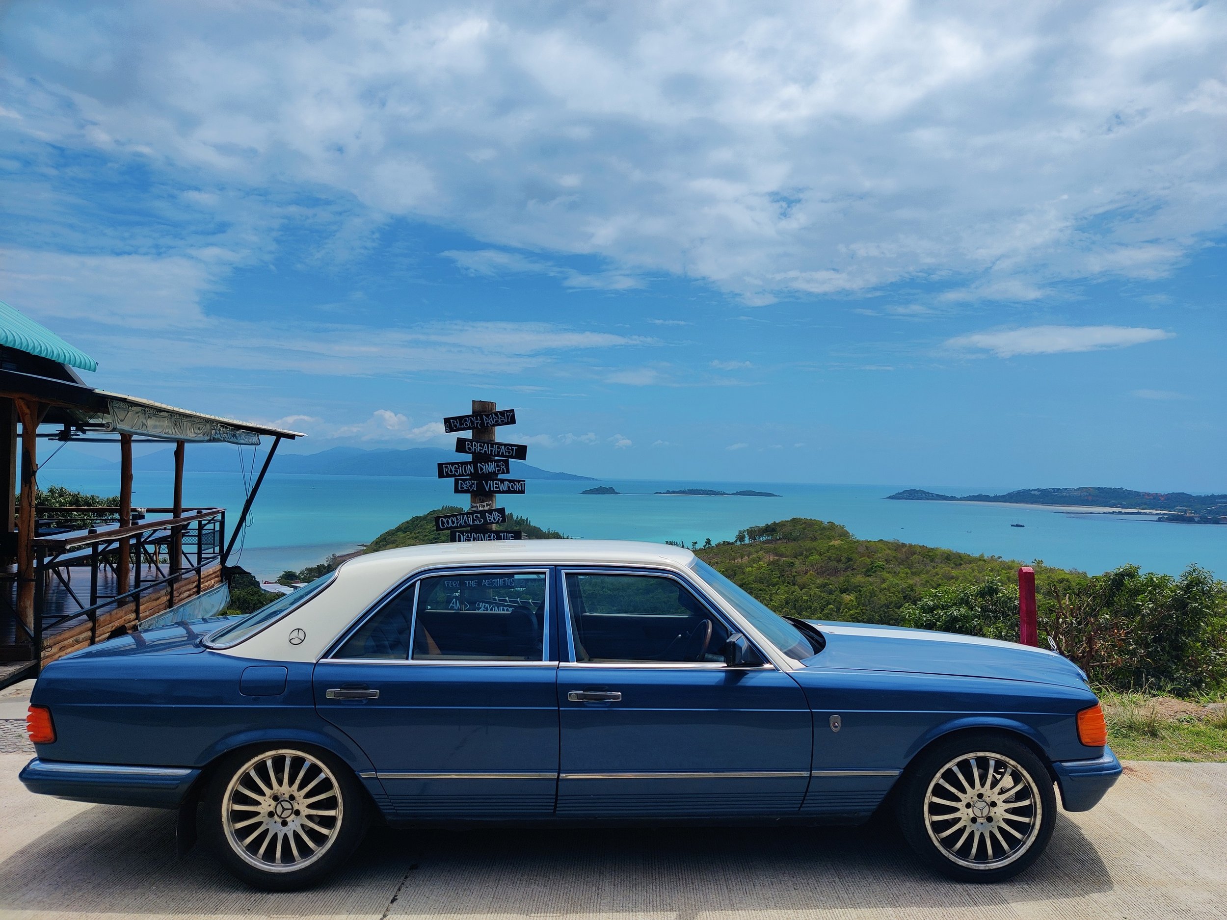 A blue vintage Mercedes-Benz car parked on a concrete surface in front of a scenic coastal view with ocean, islands, and a partly cloudy sky, with a wooden structure and signpost nearby.