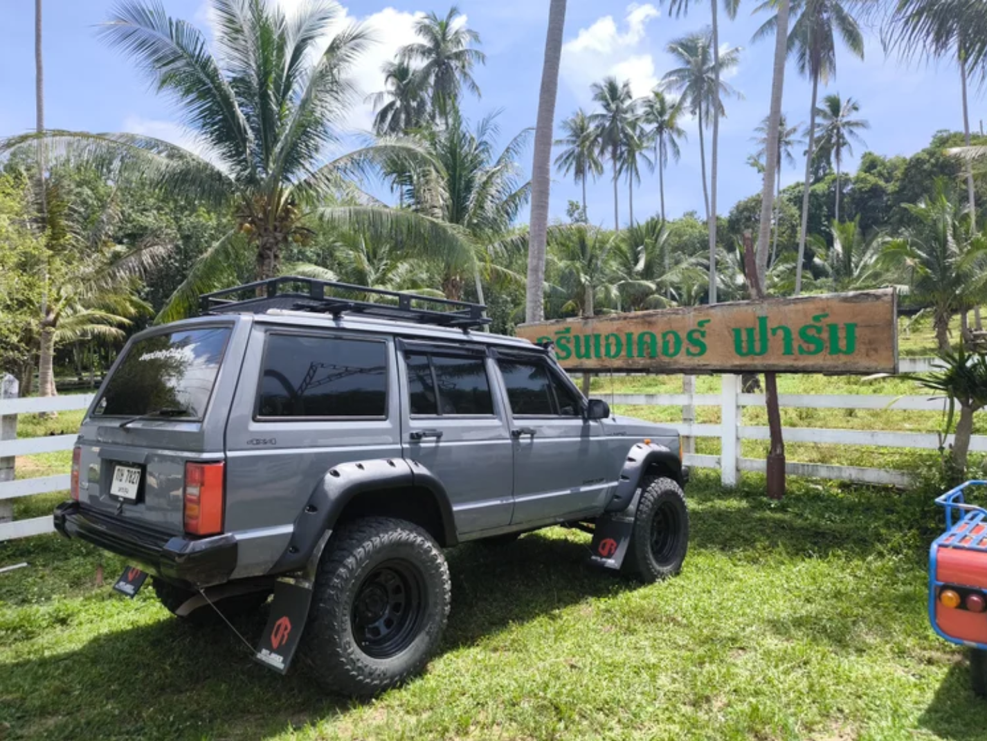 A grey off-road vehicle parked on grass in front of a wooden sign with green Thai script, surrounded by tall palm trees in a tropical setting.