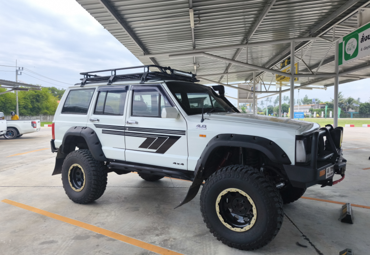 White off-road SUV with large tires and a roof rack parked under a metal roofed structure at a parking lot.