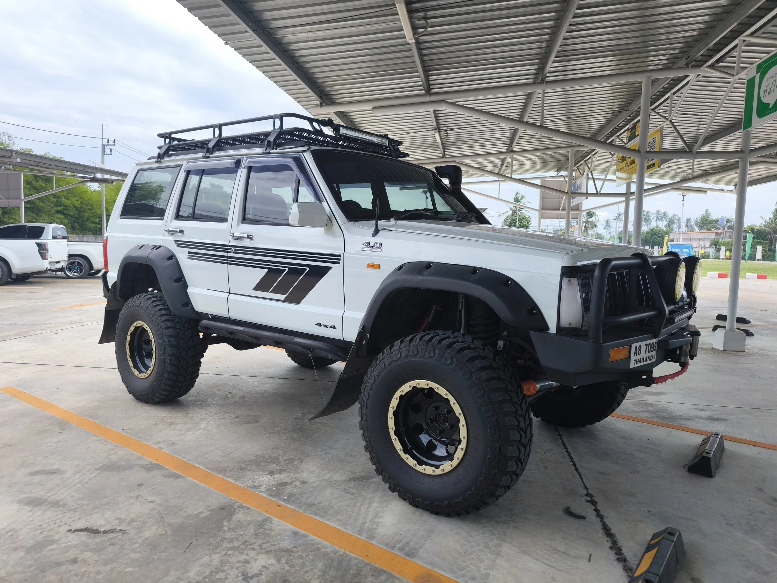 A white off-road vehicle with large tires, custom black fender flares, and a roof rack parked under a metal canopy at a parking lot.