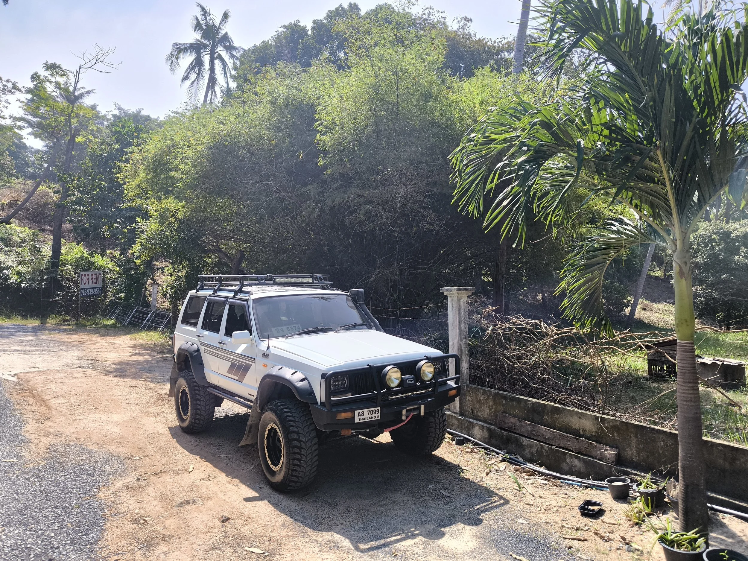 A white off-road vehicle with black front bumper and roof rack parked on a dirt area next to a large leafy tree and lush green bushes, with a clear sky above.