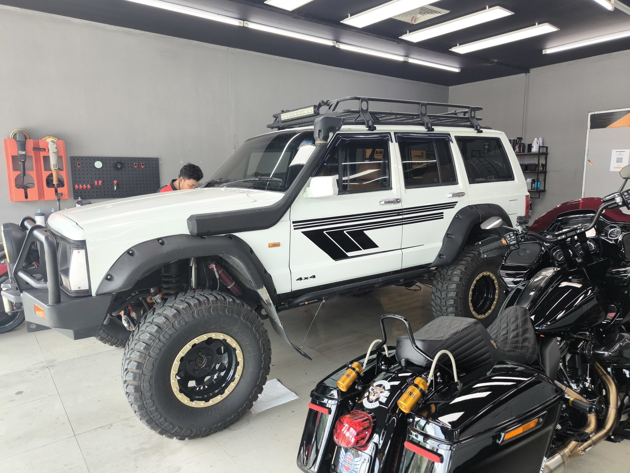 A white off-road vehicle with large tires and a rooftop cargo rack inside a garage or workshop setting, with a person working near it, motorcycle and tools visible.
