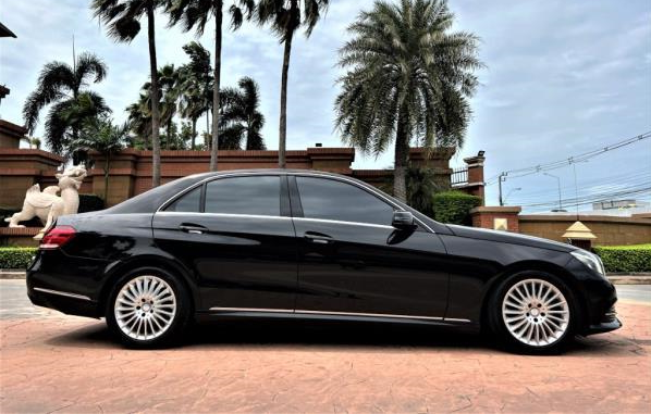 Side view of a black sedan car parked on a brick pavement, with palm trees and a brick wall in the background.