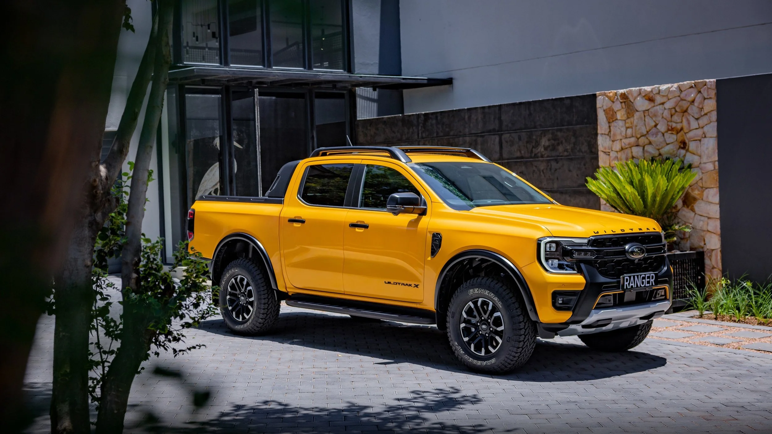 A yellow Ford Ranger Wildtrak X pickup truck parked in front of a modern building with glass windows and a stone wall, partially obscured by trees and plants.