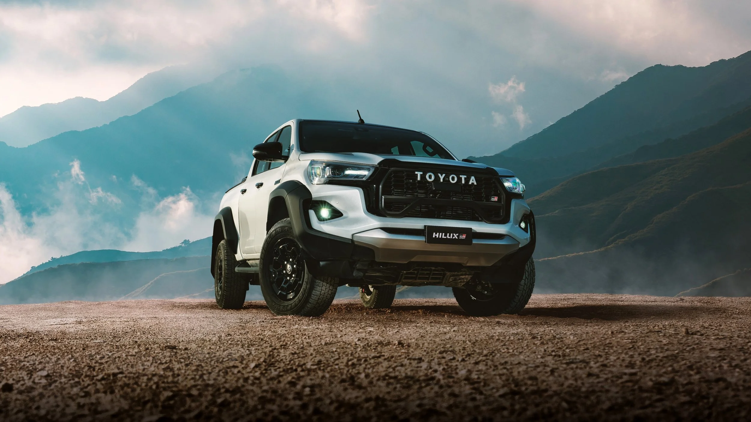 A silver Toyota Hilux GR pickup truck parked on a dirt road in front of mountains with cloudy sky.