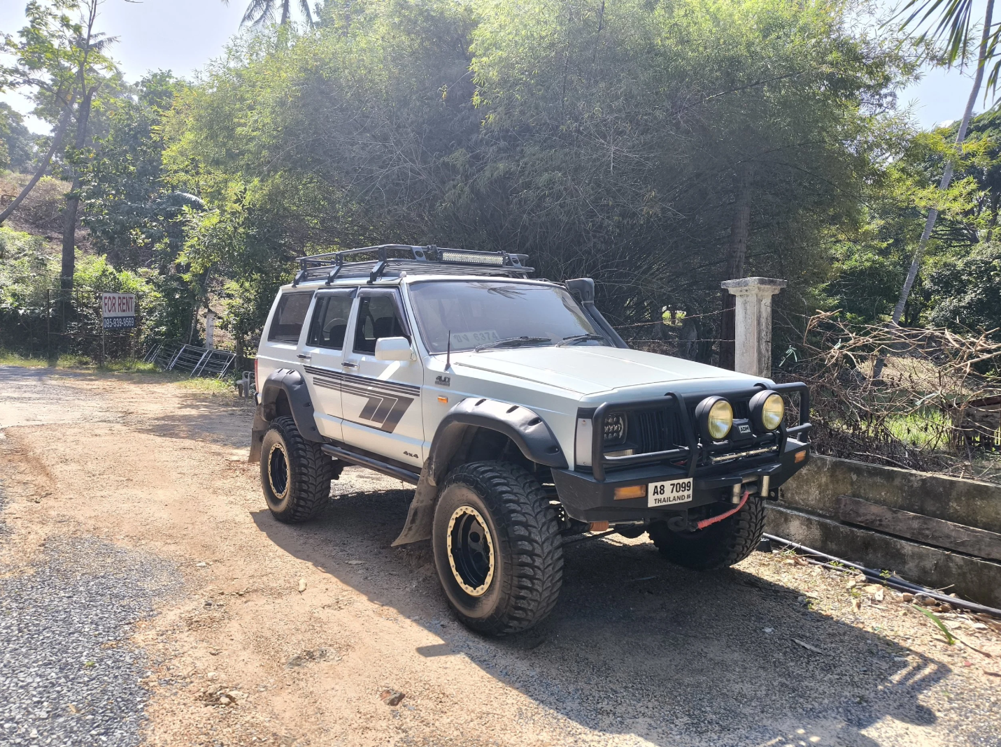 A silver off-road vehicle with large tires, a front bull bar, and roof rack parked on a dirt road near trees and a fence with a 'For Rent' sign in the background.