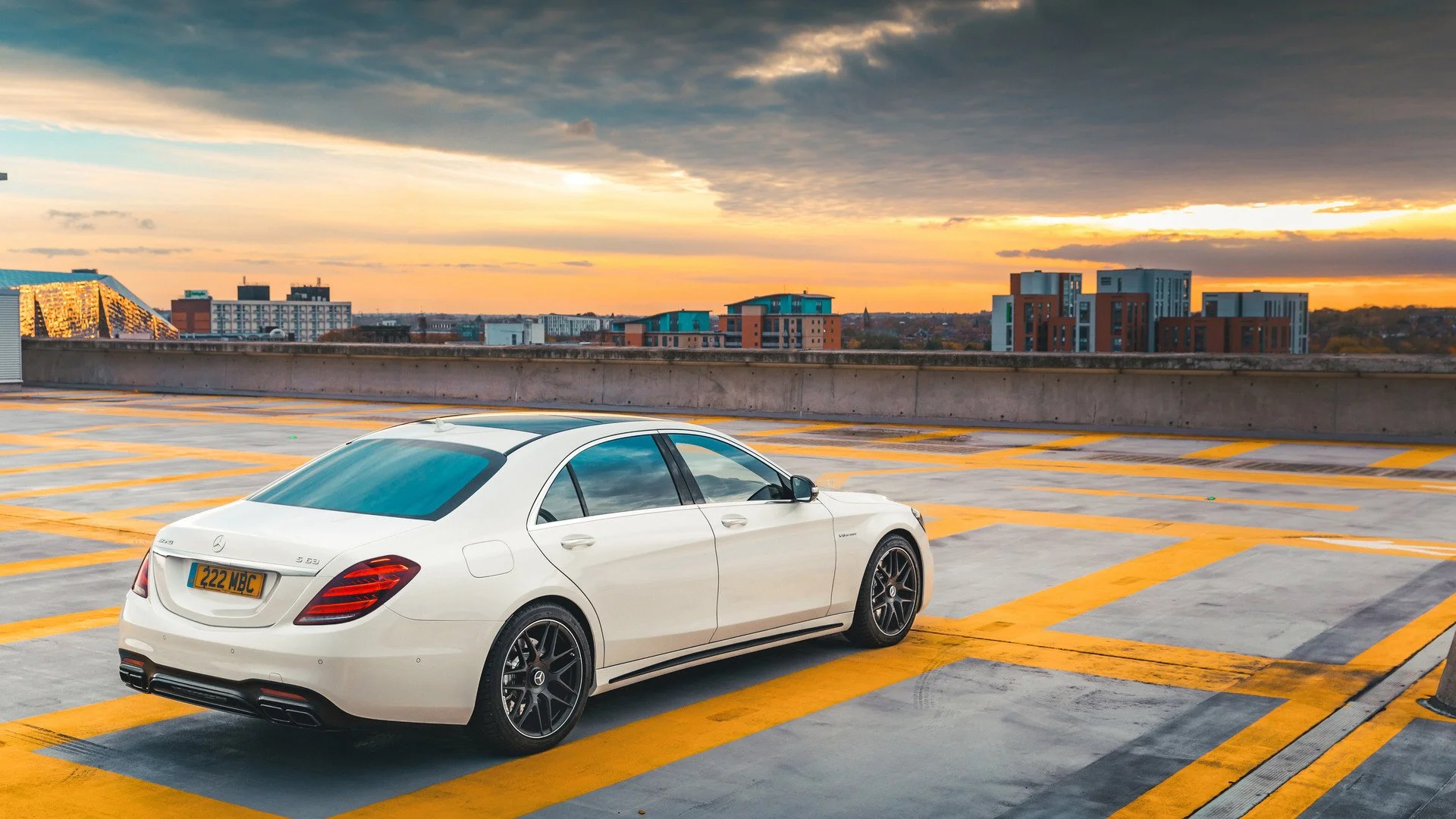 A white luxury Mercedes-Benz parked on the rooftop of a parking garage during sunset. The city skyline with various buildings is visible in the background under a partly cloudy sky.