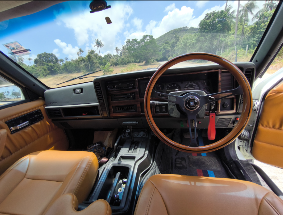 Interior view of a Jeep vehicle showing the dashboard, steering wheel, and tan leather seats, with a scenic tropical landscape with trees and mountains visible through the windshield.