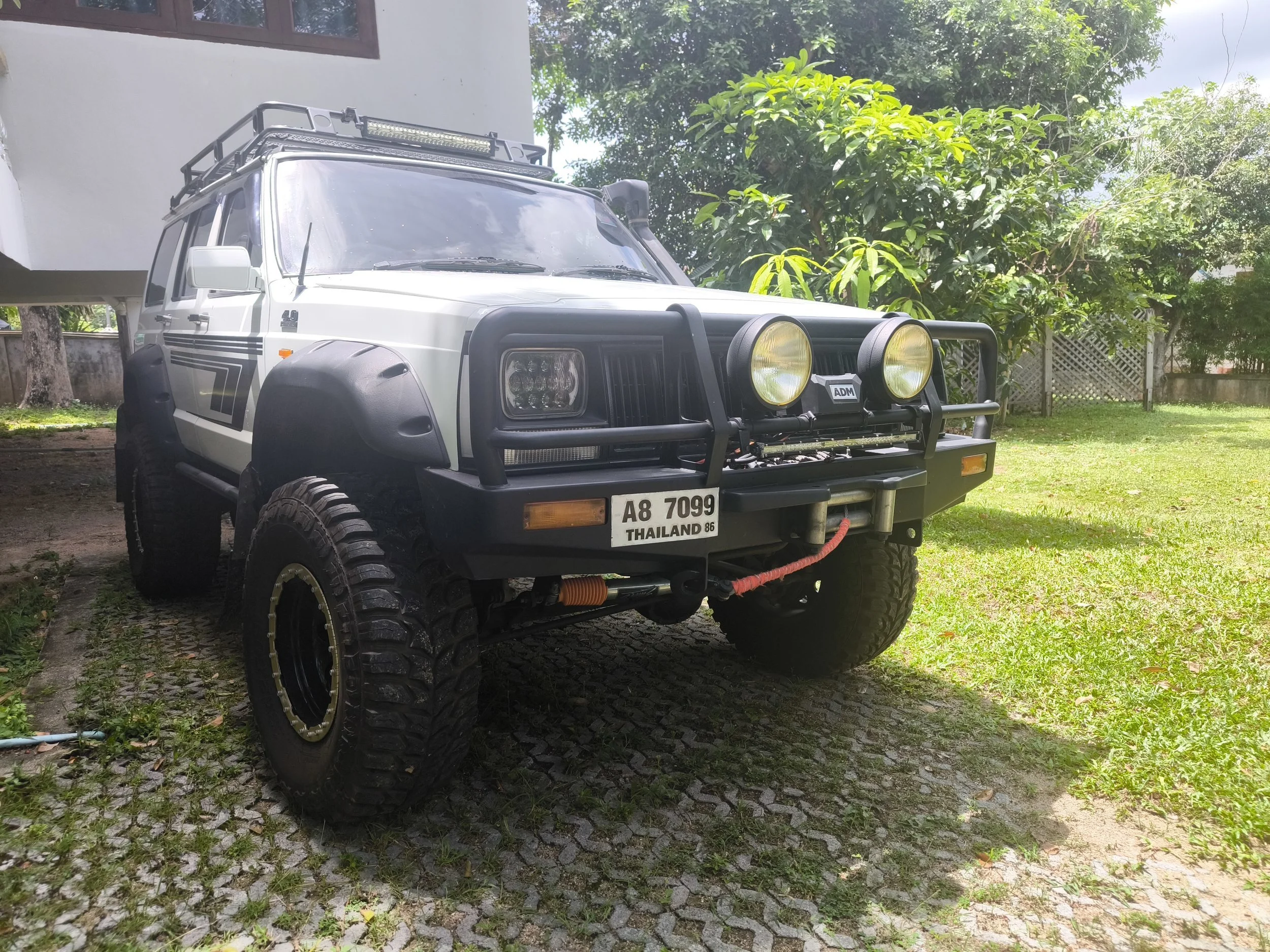 A white off-road vehicle parked on a cobblestone driveway, surrounded by greenery and trees, with a house in the background.