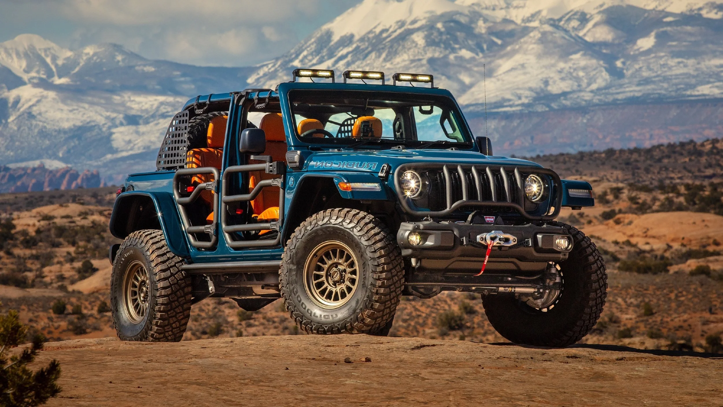 A blue off-road vehicle with large tires and orange seats parked on desert terrain with mountains in the background.