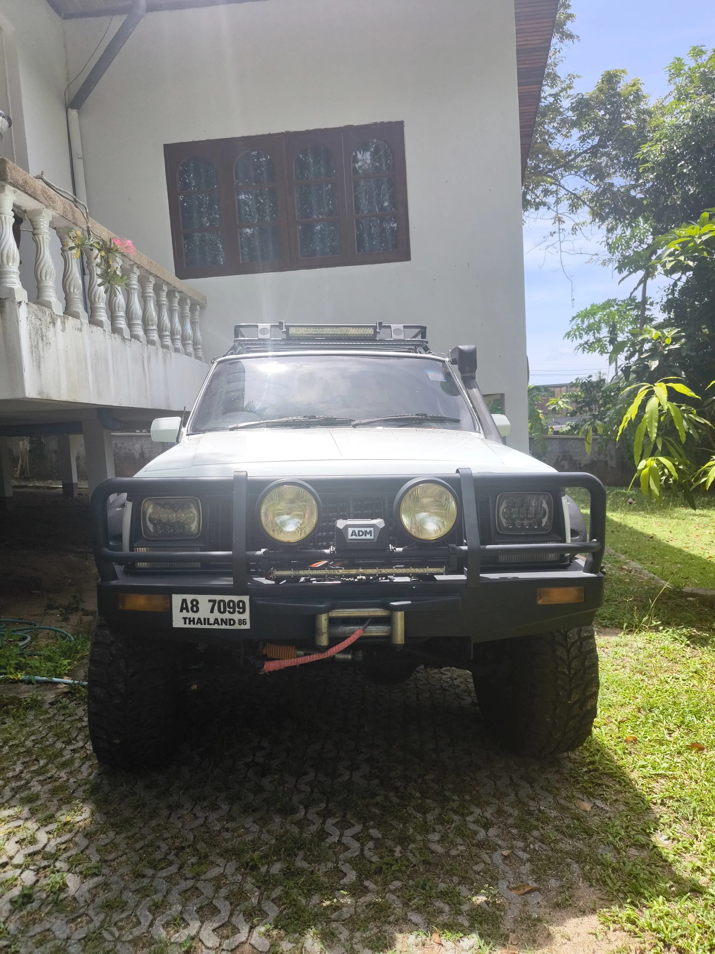 A white off-road vehicle with a front bumper guard, large tires, and a roof rack parked in front of a building. The vehicle has a license plate reading 'A8 7099' from Thailand, and a sticker with 'ADM' on the front. The building has a window with woo
