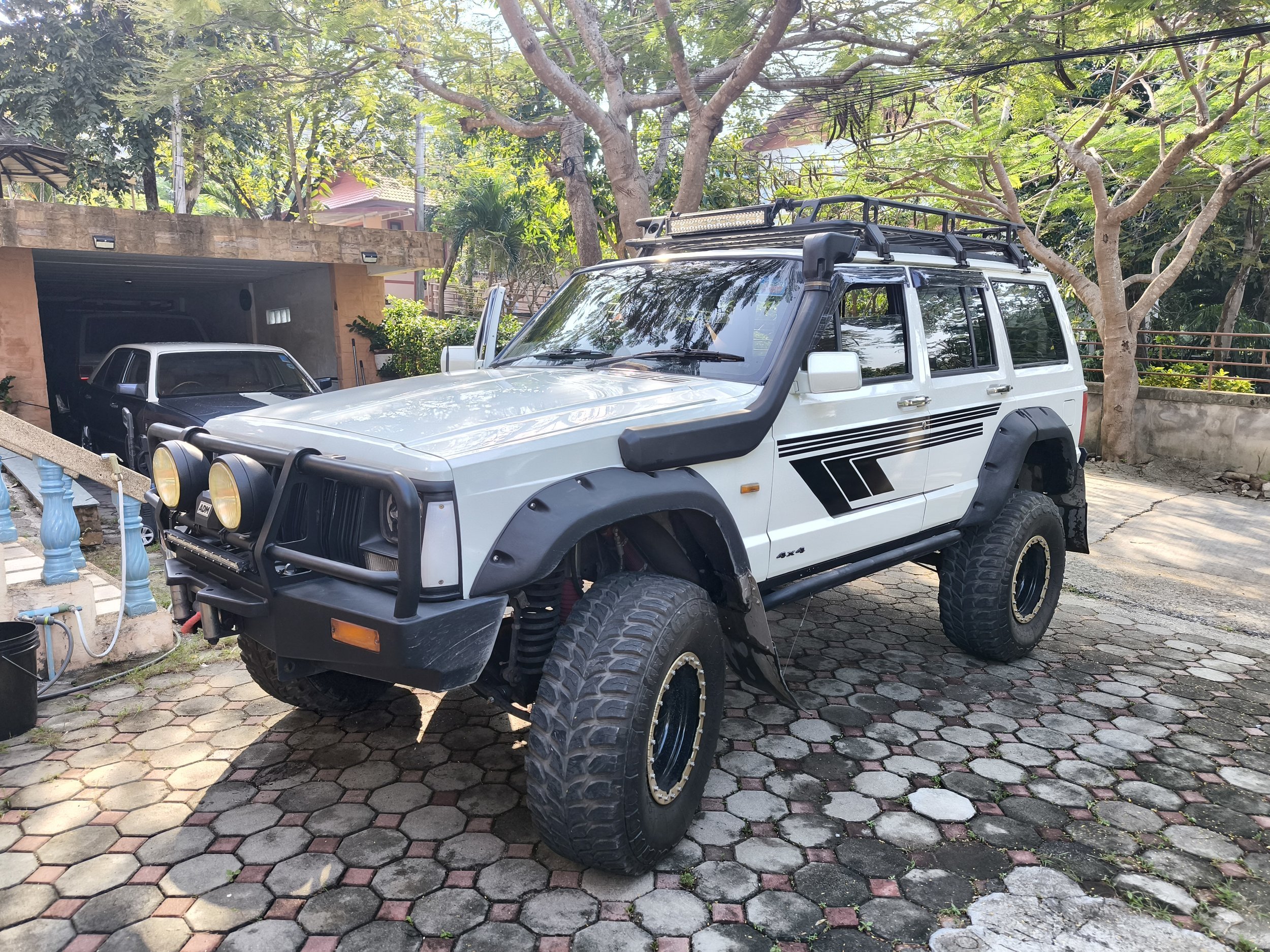 A white off-road vehicle with large tires and a roof rack parked in a driveway under trees, with a black car parked in a garage in the background.