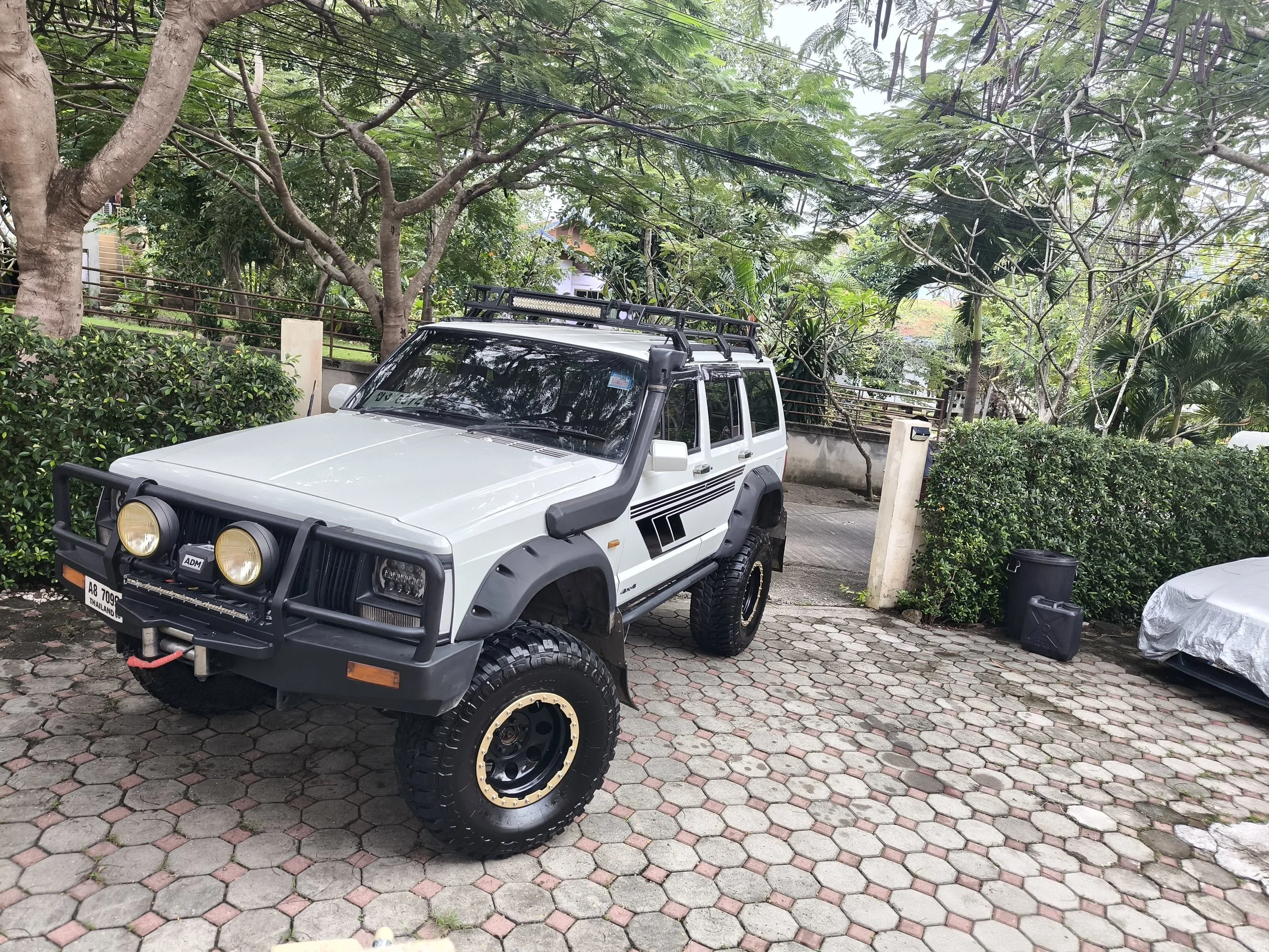 A white off-road vehicle with black accents and round headlights parked on a paved driveway surrounded by lush green trees and bushes.