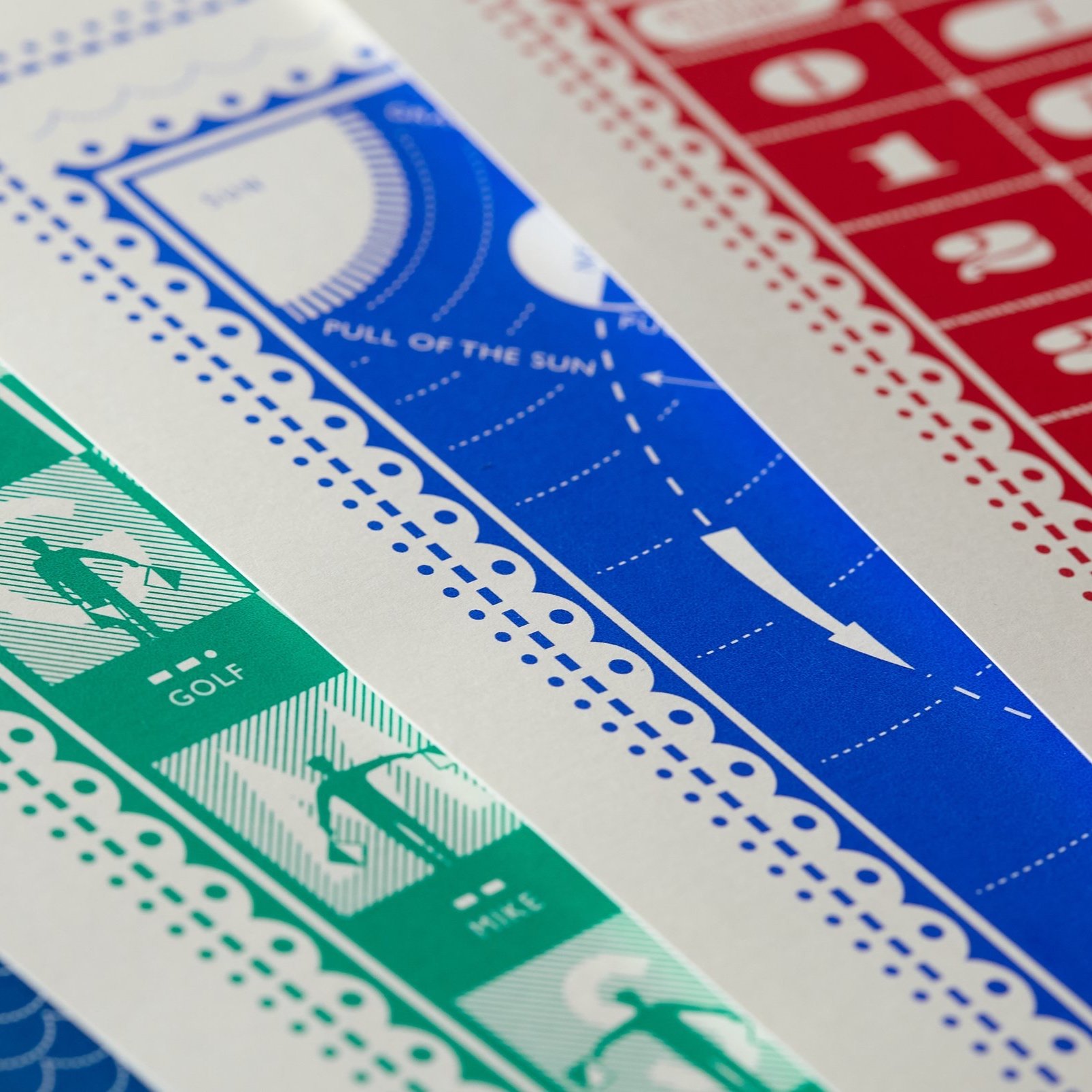 Close-up of a color-coded printed design with blue, red, and green sections. The green section has silhouettes of figures labeled "GOLF" and "HIKE." The blue section features text "FULL OF THE SUN" with arrows and dotted lines. The red section contai